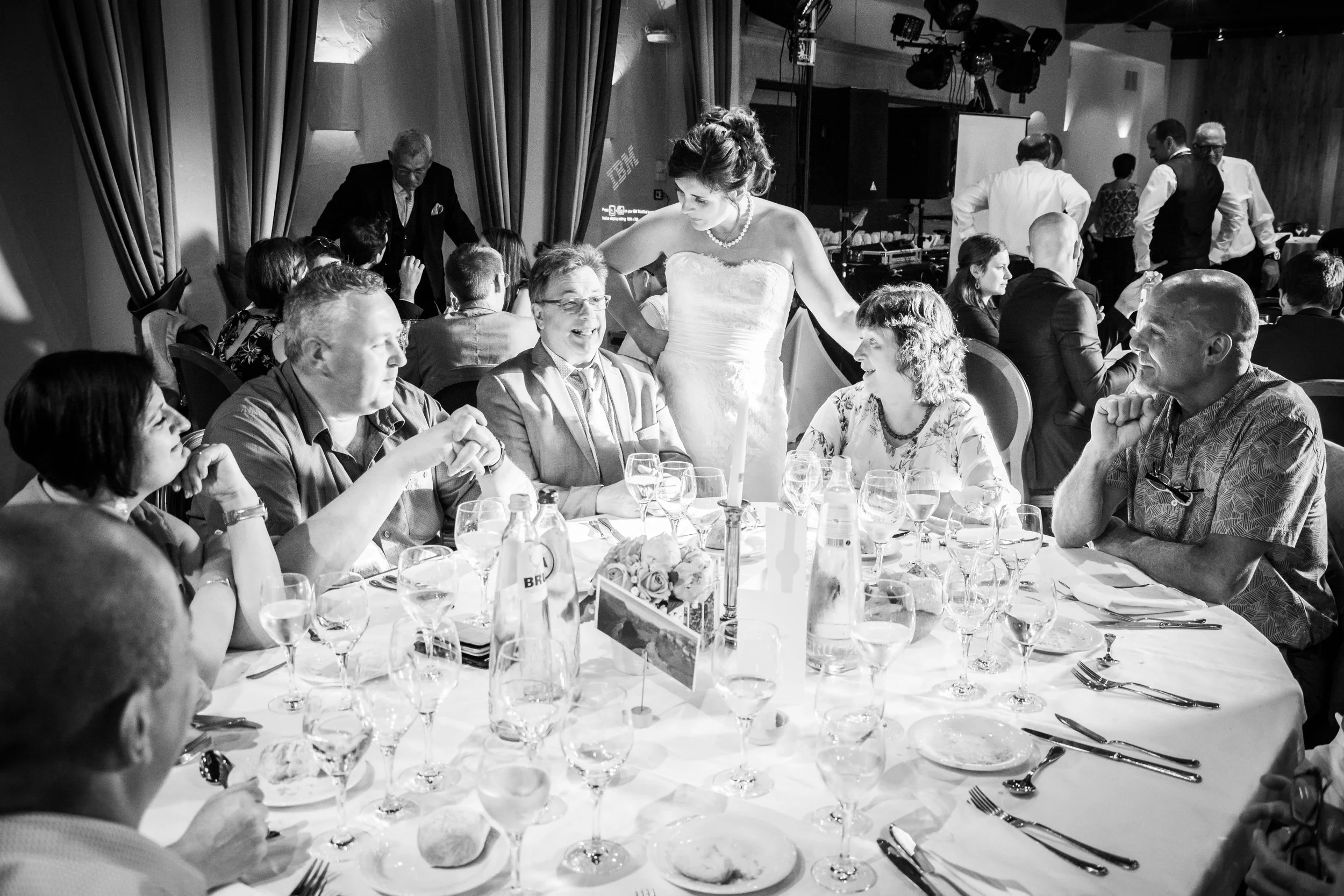Op een feestzaal zitten gasten aan een ronde tafel, midden in gesprek en genieten. Een vrouw in een witte trouwjurk staat tussen de gasten en glimlacht.