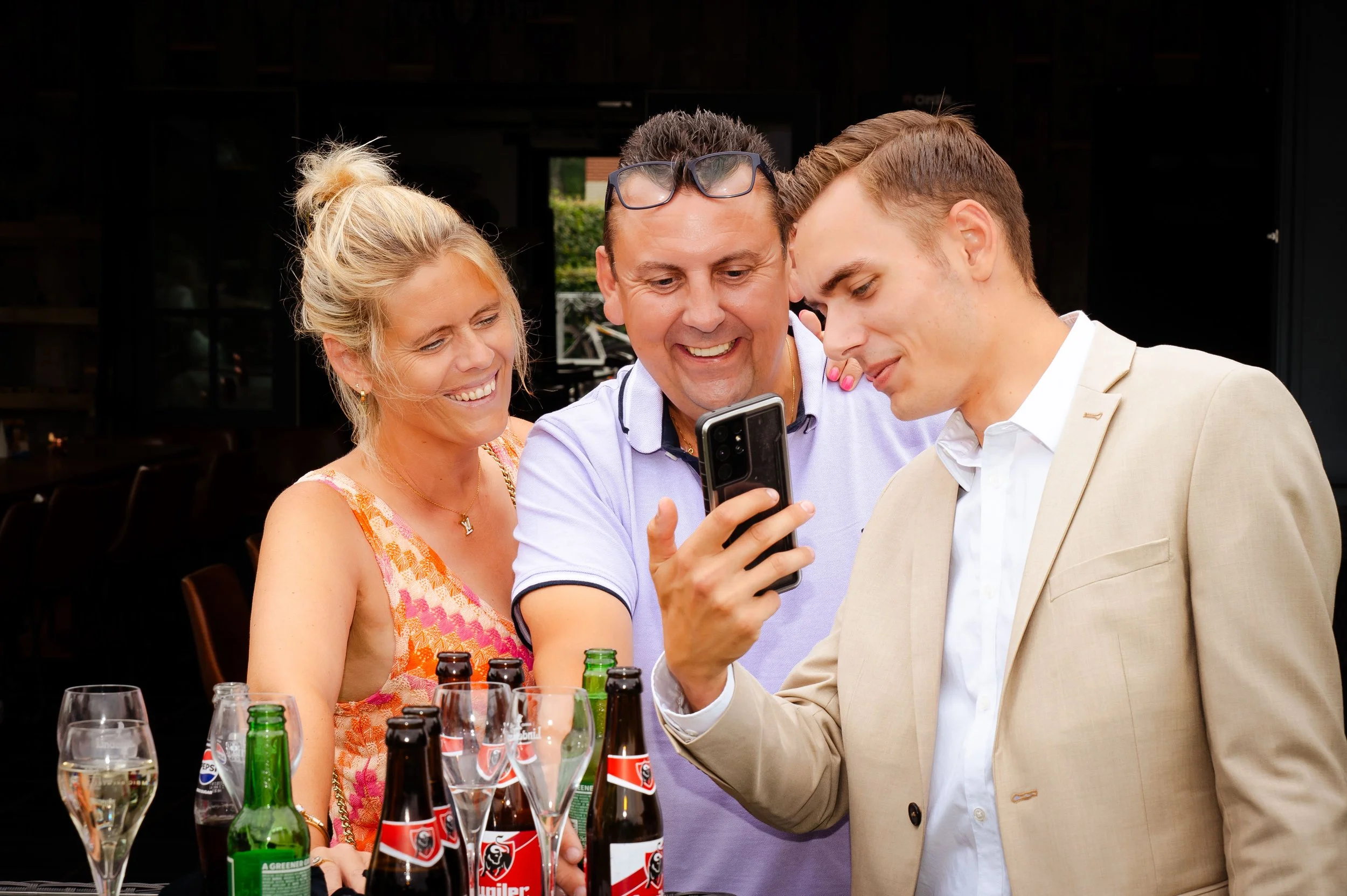 Drie mensen die lachend naar een smartphone kijken tijdens een bijeenkomst buitenshuis, met drankjes en bierflesjes op tafel.