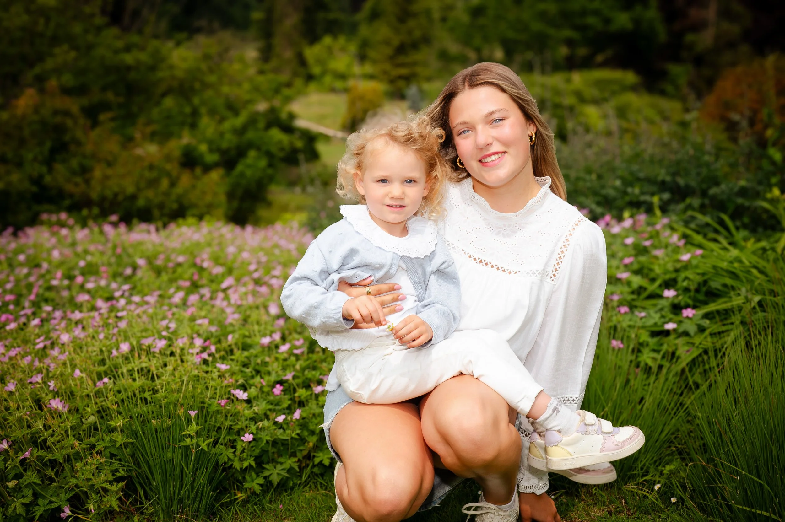 Vrouw en klein meisje zittend op grasveld met bloemen, achtergrond van groen bos en land.