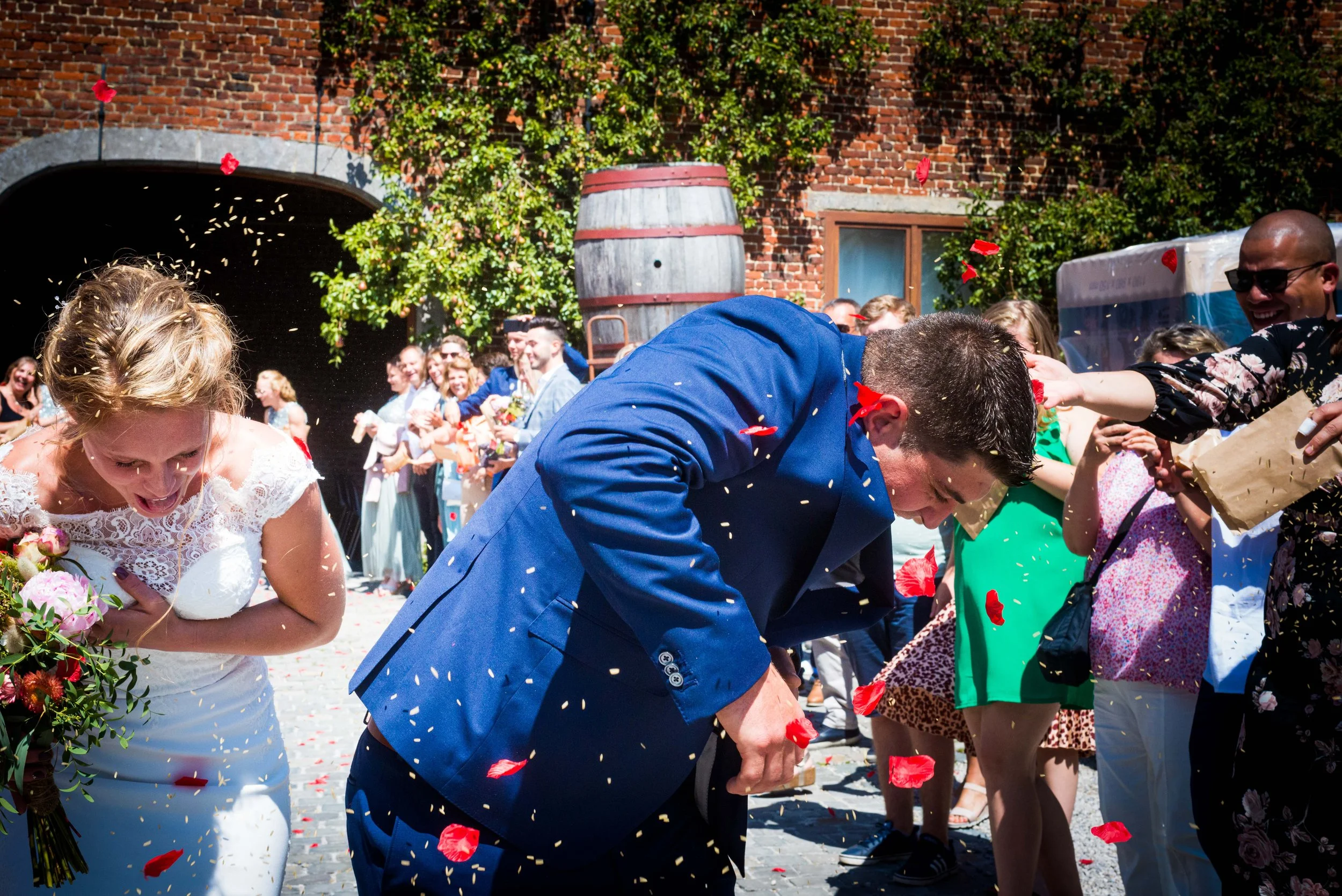 Bruid en bruidegom die zich bukken terwijl confetti wordt geworpen tijdens een feestelijke gelegenheid, met gasten die toekijken en lachen op een straat met bakstenen muur en bomen op de achtergrond.
