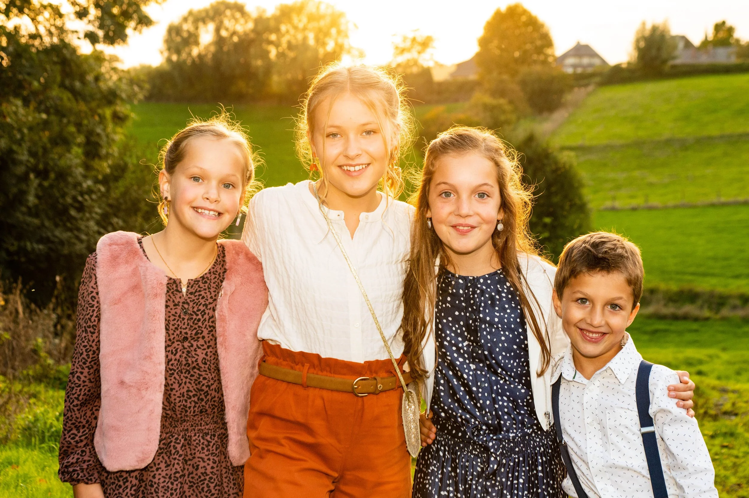 Vijf kinderen staan samen in een veld bij zonsondergang, lachend en gekleed in casual kleding.