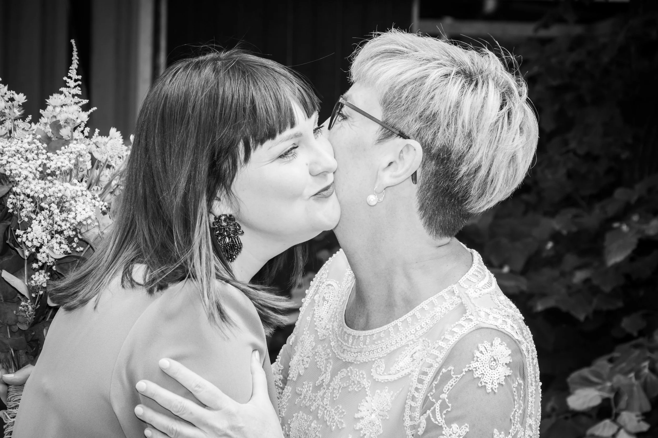 Twee vrouwen die elkaar kussen, nabij bloemen en groene planten, in zwart-wit