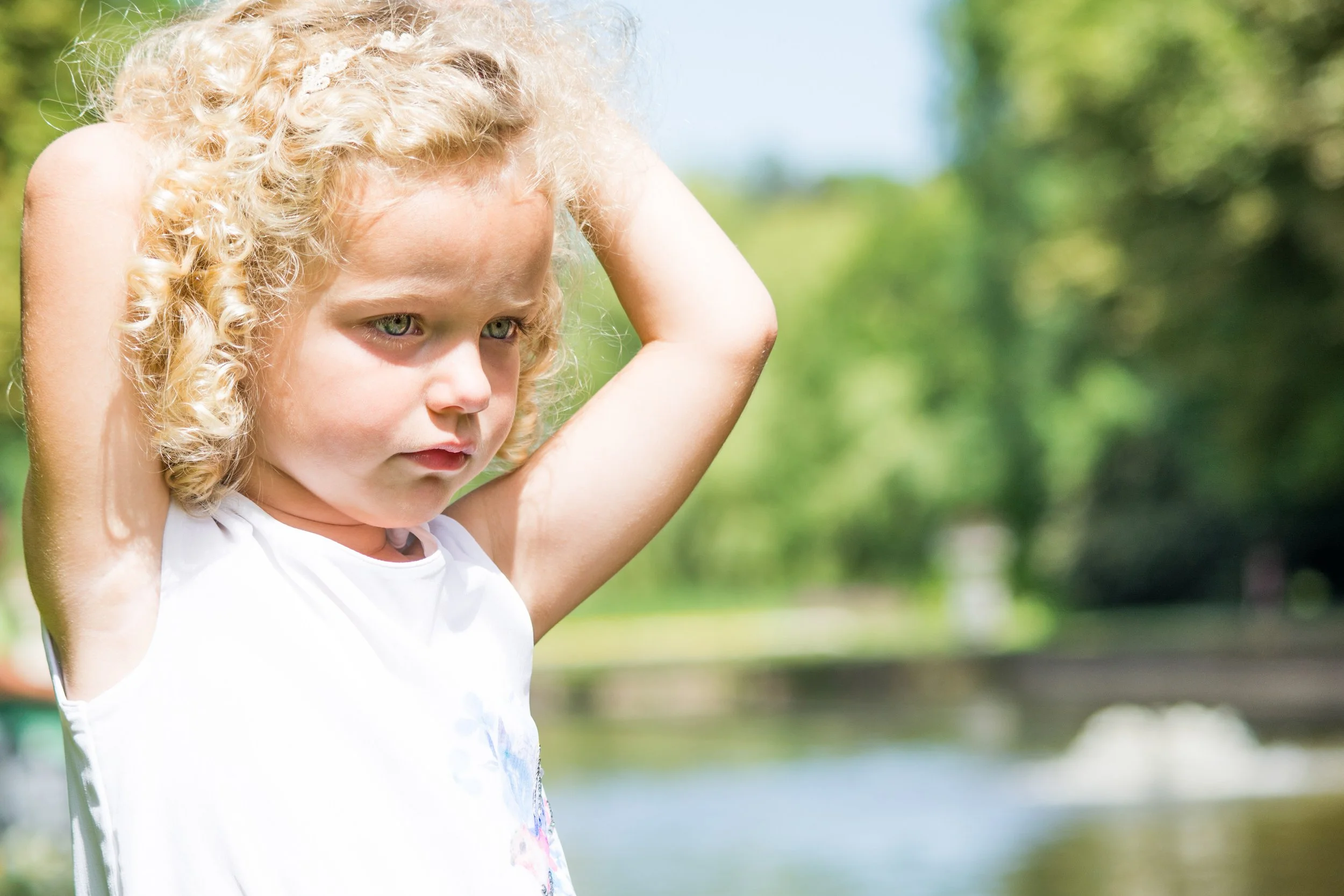 Een jonge blondharige meisje met krullend haar zit buiten, met haar handen achter haar hoofd, in een groene natuurlijke omgeving.