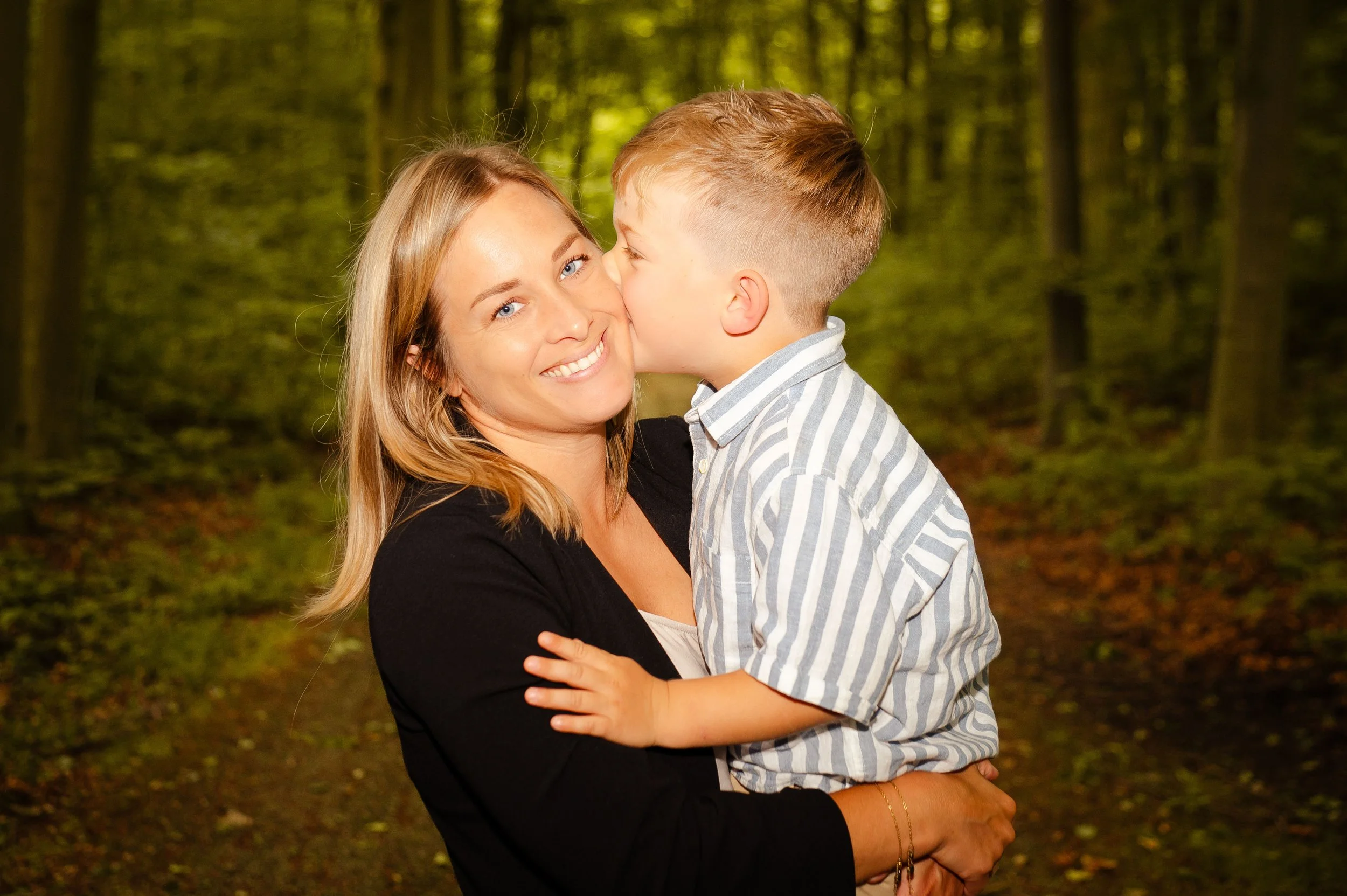 Een vrouw houdt een jonge jongen in haar armen, ze kussen elkaar in een bos met groene bladeren en bomen.