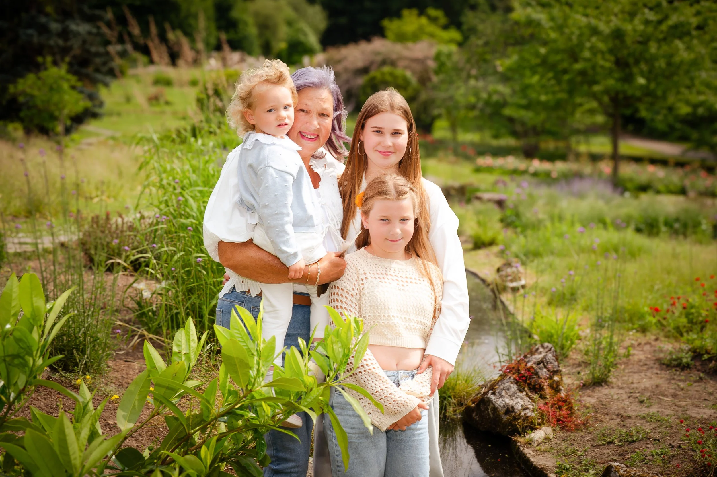 Vrouw met drie jonge meisjes poseren in een tuin met groene planten, bloemen en een kleine waterstroom, allemaal lachend en gekleed in lichte kleding.