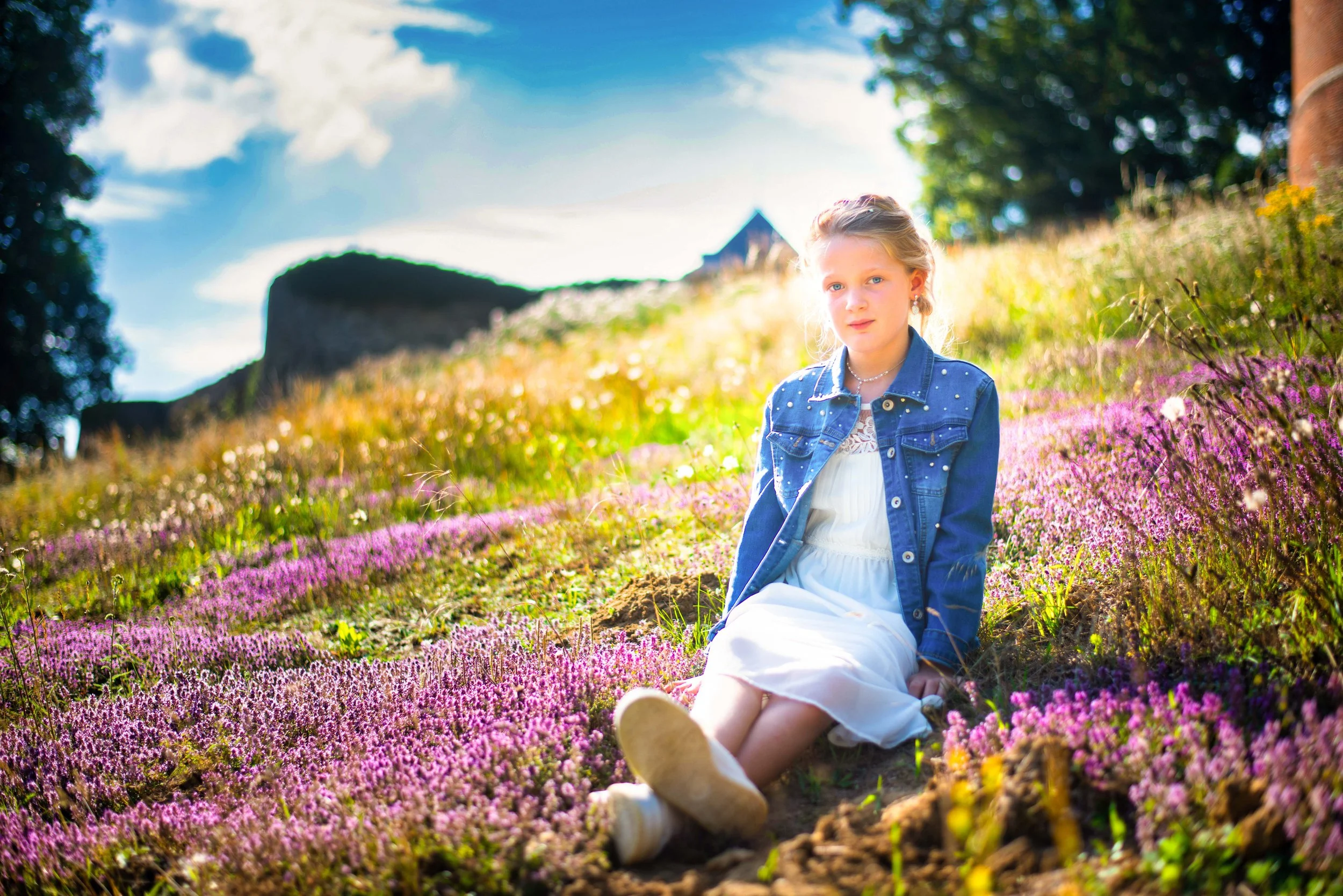 Een jonge meisje zit op een bloemenveld met paarse en roze bloemen, gekleed in een witte jurk en een blauwe spijkerjack, met een bos in de achtergrond onder een blauwe lucht met wit wolken, tijdens een zonnige dag.