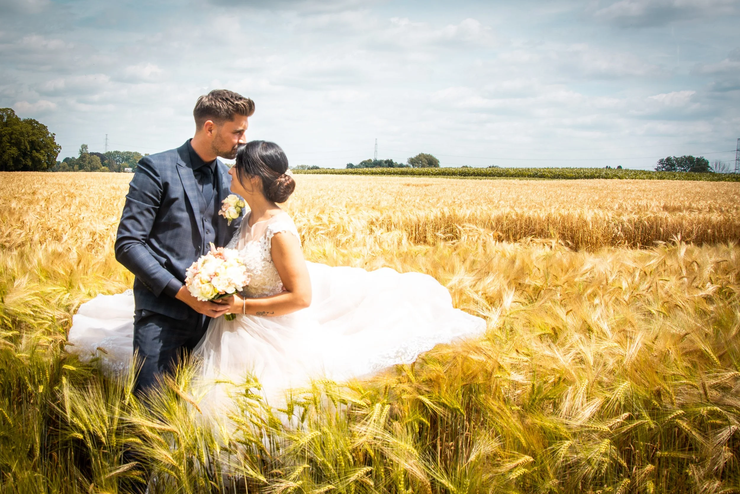 Een trouwpaar staat in een veld met gouden gerst, de man in een donkerblauw pak en de vrouw in een witte trouwjurk, ze houden een boeket bloemen vast en naderen elkaar intiem.