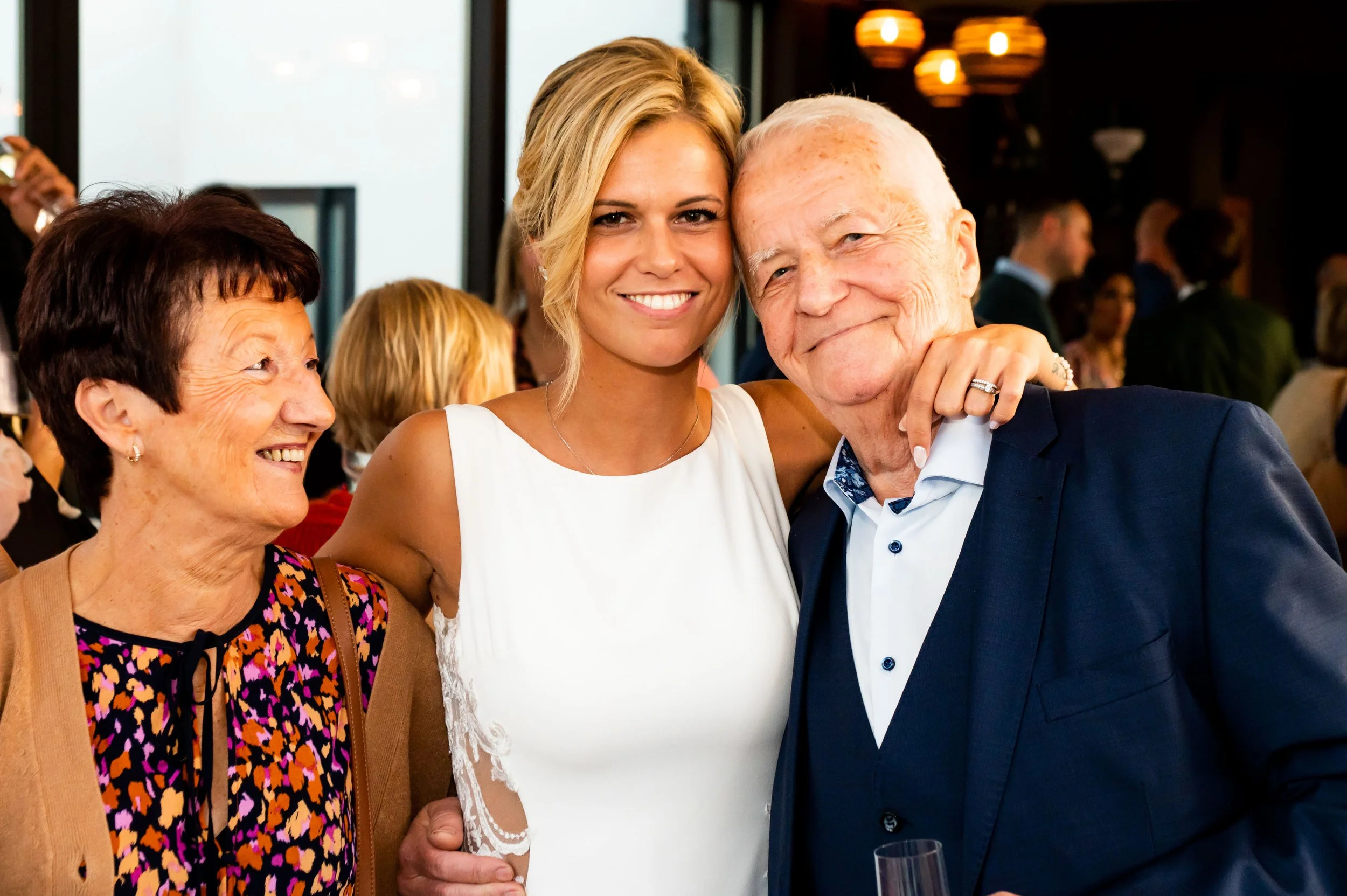 Drie mensen glimlachend samen op een feest, een oudere vrouw, een jonge vrouw en een oudere man, omringd door andere gasten.