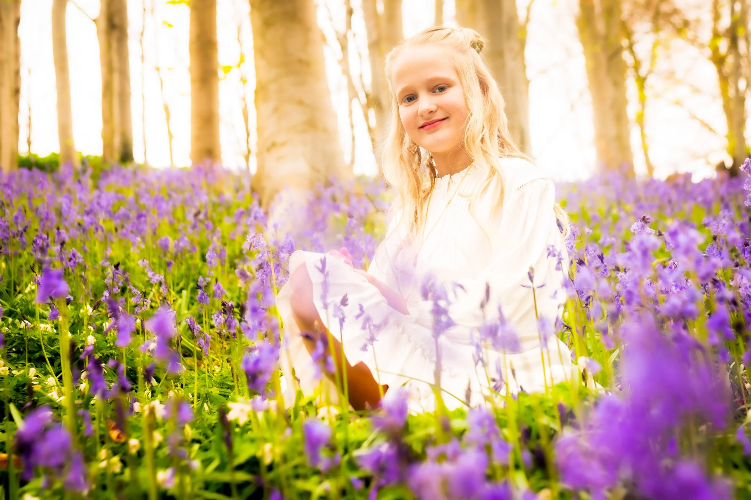 Een meisje zit tussen paarse bloemen in een bos met bomen op de achtergrond, onder een warme zon.