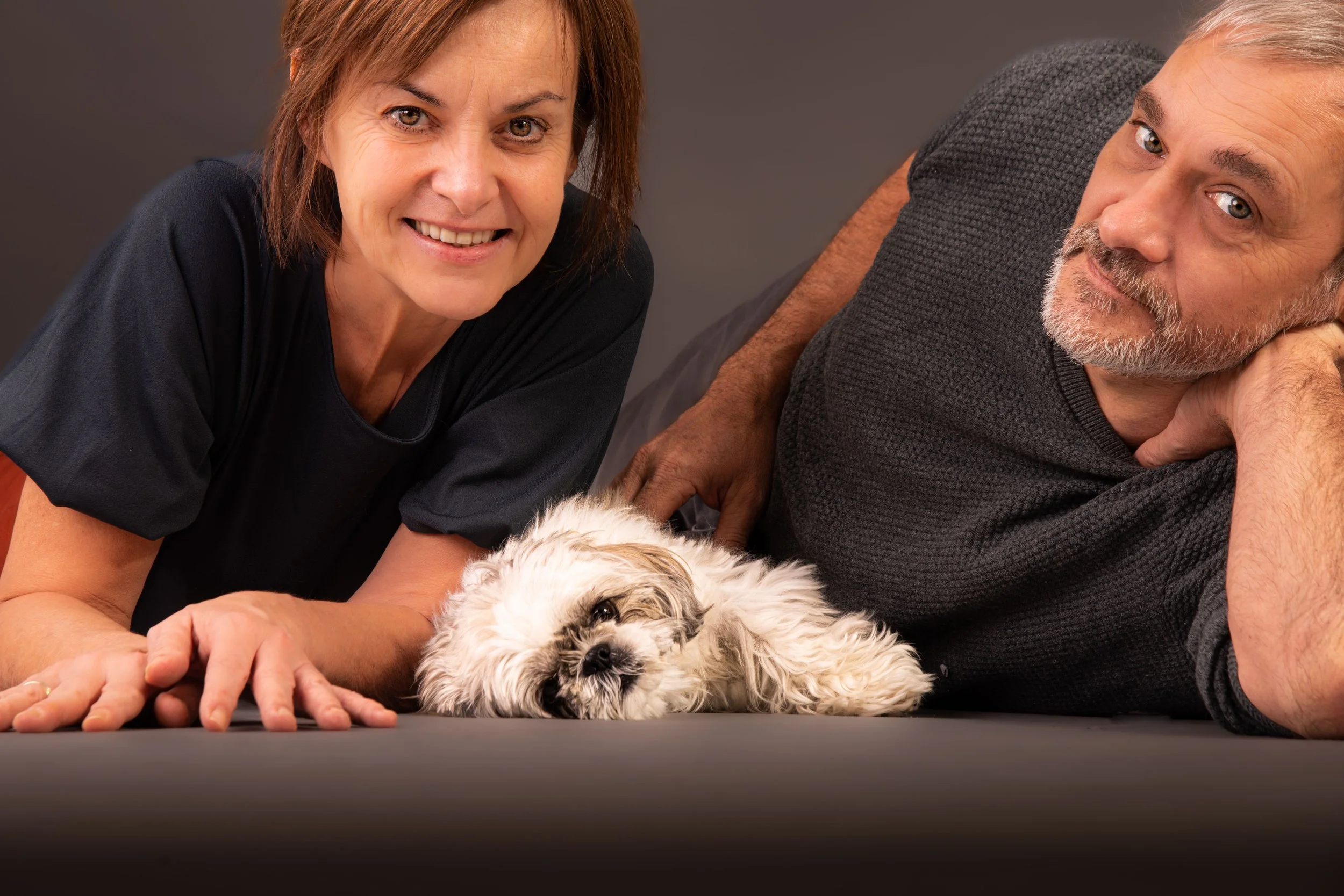 Een vrouw en een man liggen naast een slapende kleine witte en bruine hond op een grijze achtergrond.