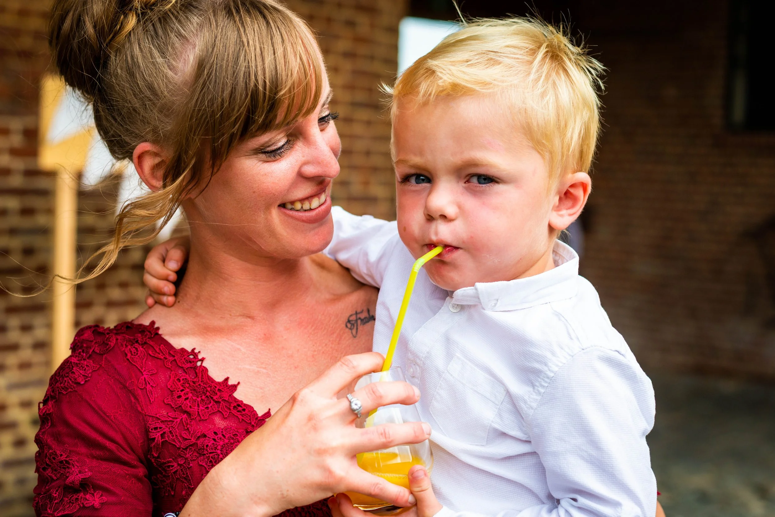 Een vrouw in een rode jurk houdt een jonge jongen vast terwijl hij aan een drankje met een gele rietje nippt. Ze glimlacht en kijken naar elkaar.