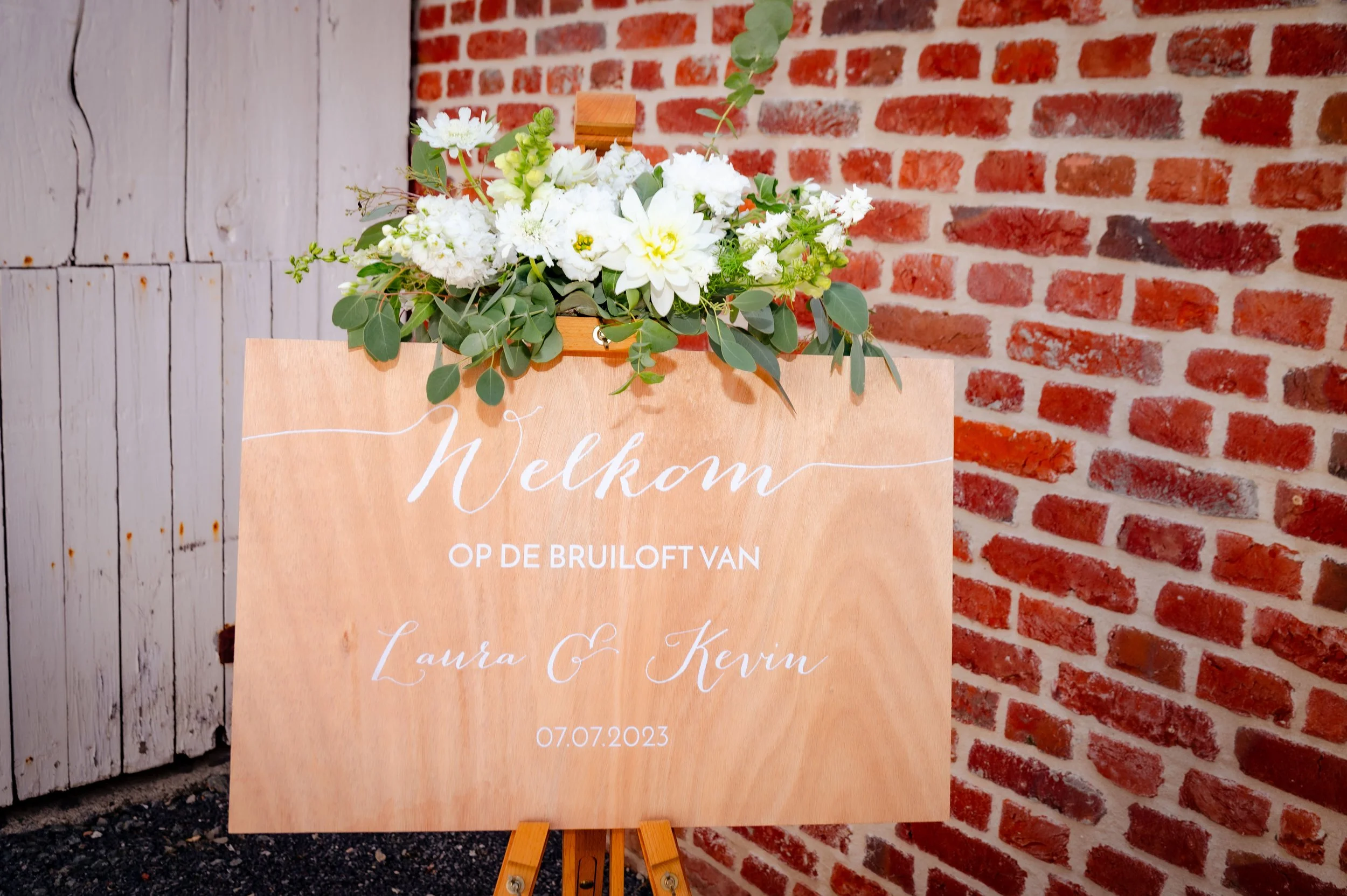 Welkomstbord met bloemen, houten bord met witte tekst voor bruiloft op 07.07.2023, bloemen en groen boven het bord, bakstenen muur en houten deur op achtergrond.