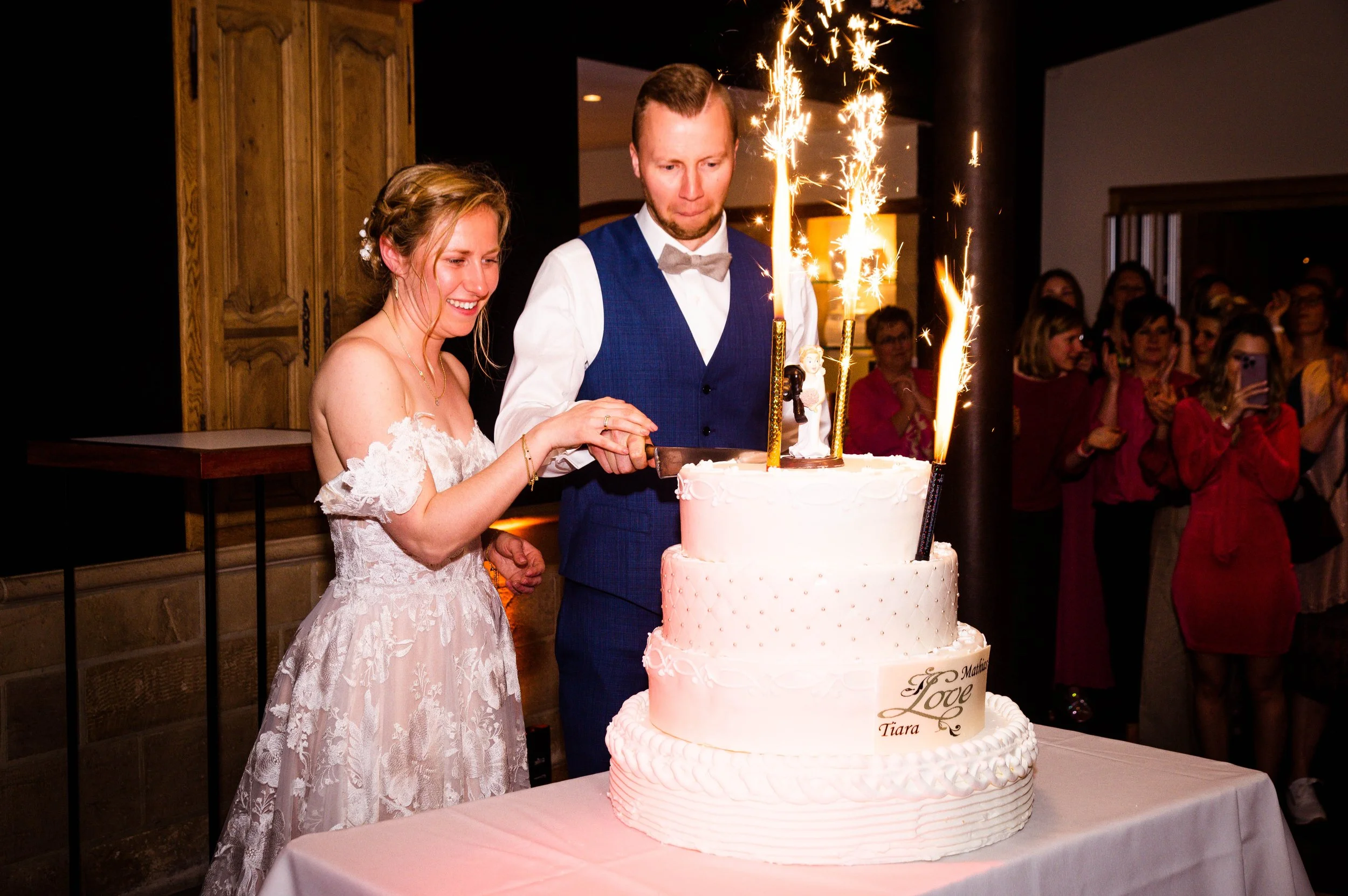 Een bruid en bruidegom snijden samen een grote, witte bruidstaart met sparklers erop tijdens hun huwelijk. De bruid draagt een witte kanten jurk en lacht, terwijl de bruidegom in een blauwe vest en wit overhemd is.