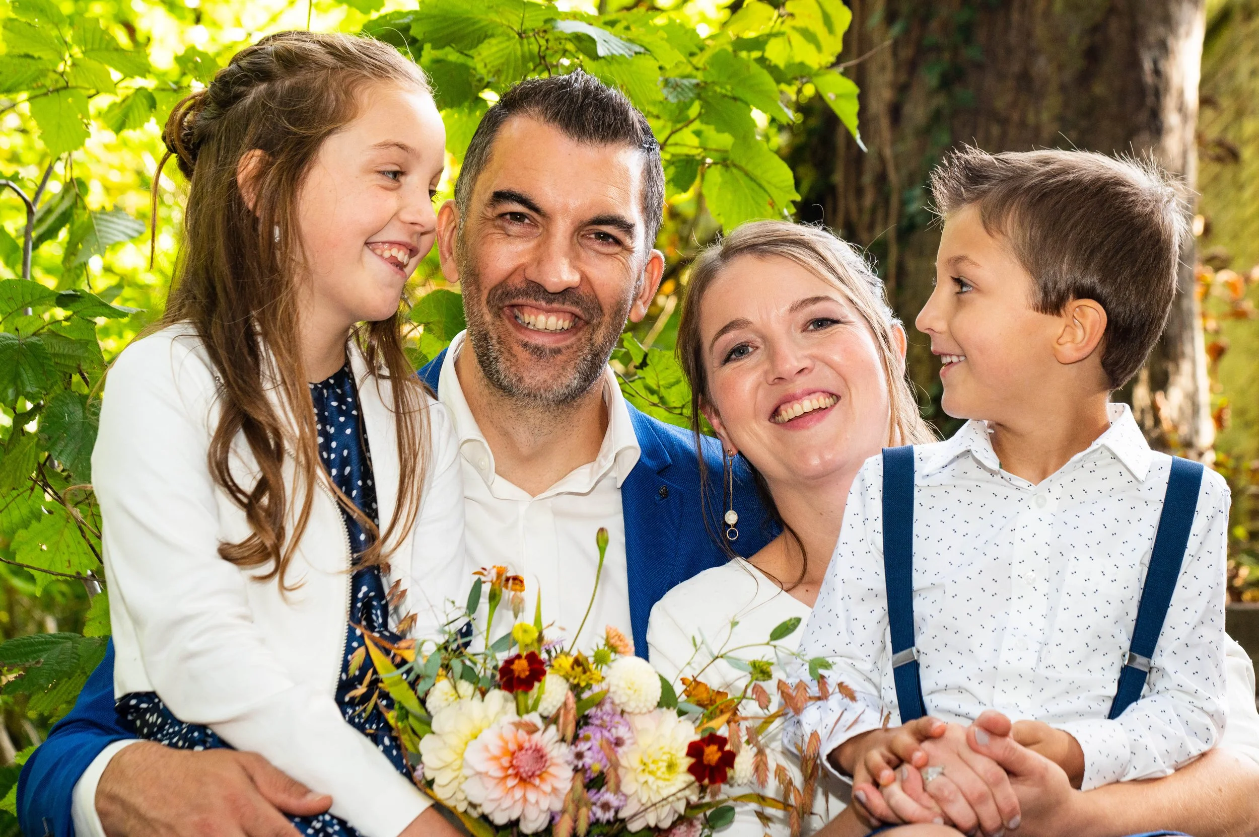 Gezicht van een gezin van vijf mensen dat buiten in de natuur staat, omringd door groene bladeren, met twee jonge kinderen en een baby, allen lachend en genietend.