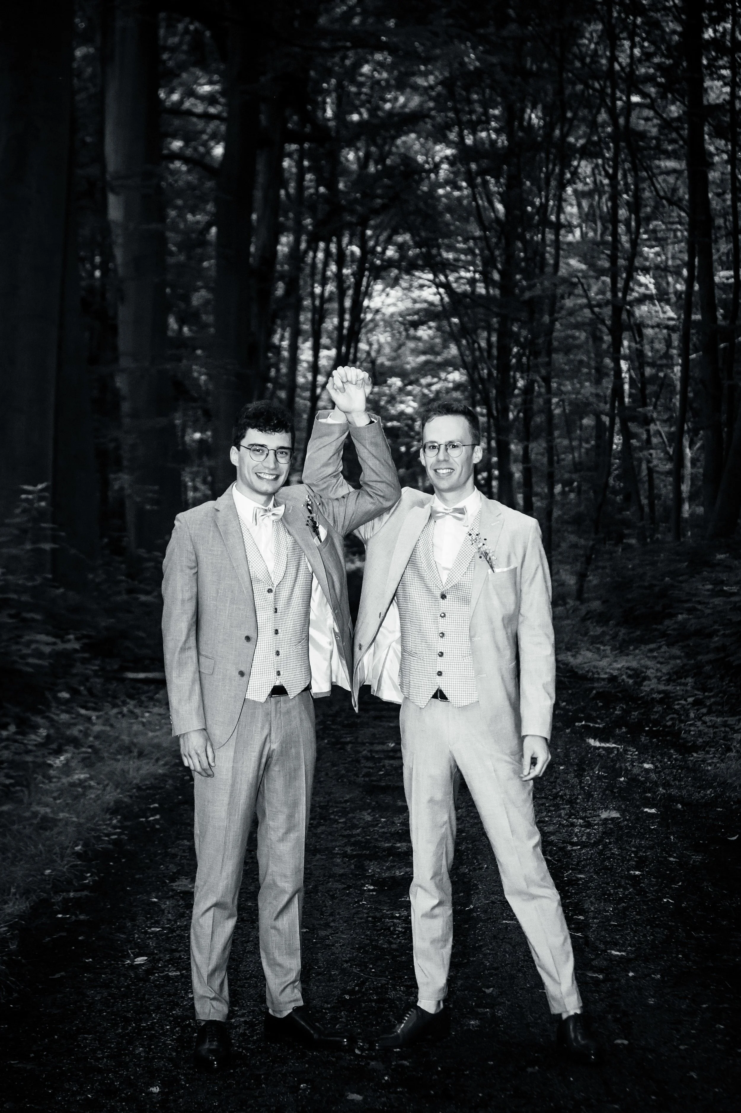 Twee mannen in trouwkleding poseren in een bos, één van hen wordt omhoog gehouden en beiden lachen.