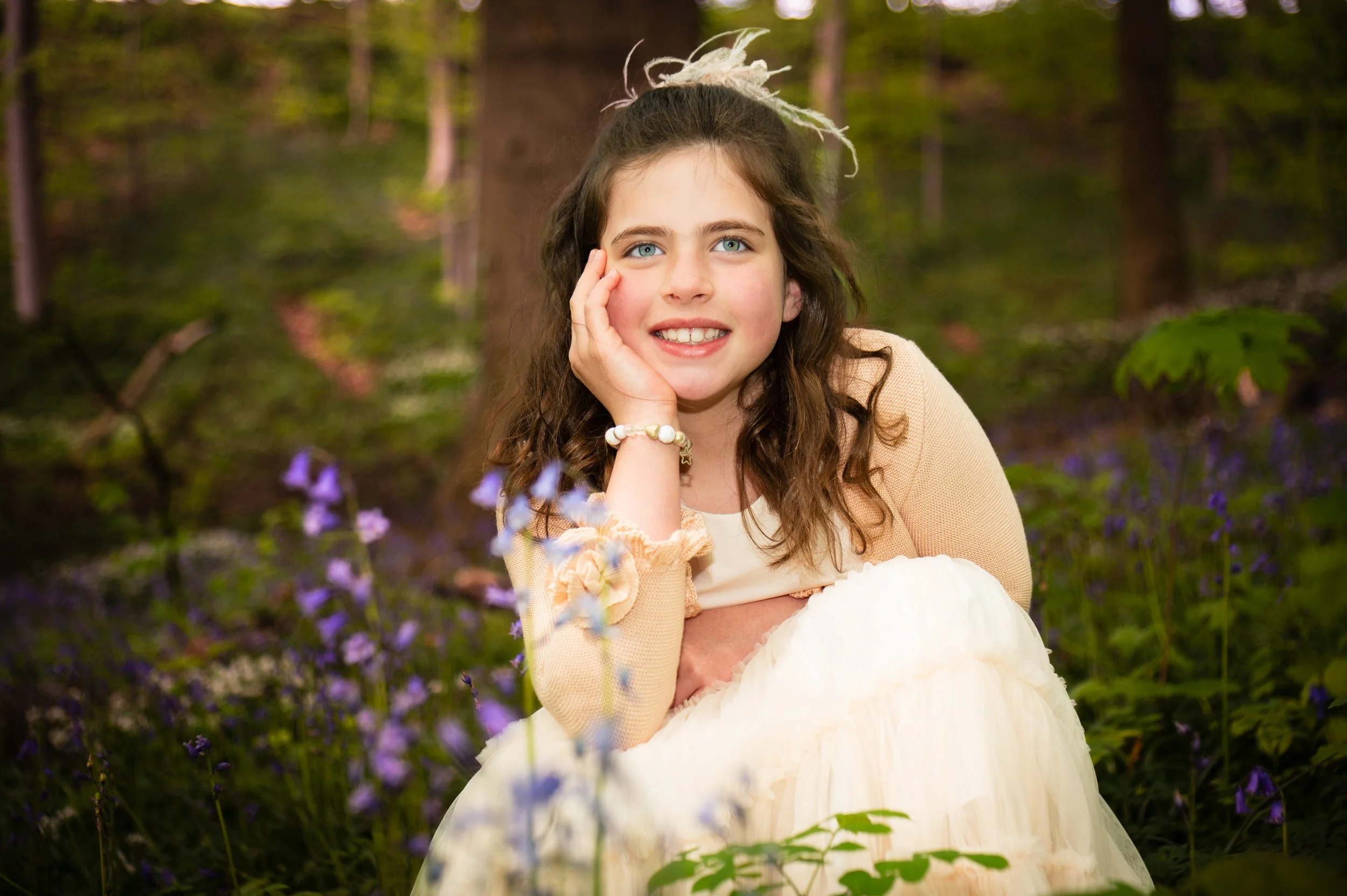 Een jonge vrouw met lange, golvende bruin haar en blauwe ogen, zittend in een bos met bloemen, glimlachend met haar hoofd rustend op haar hand.