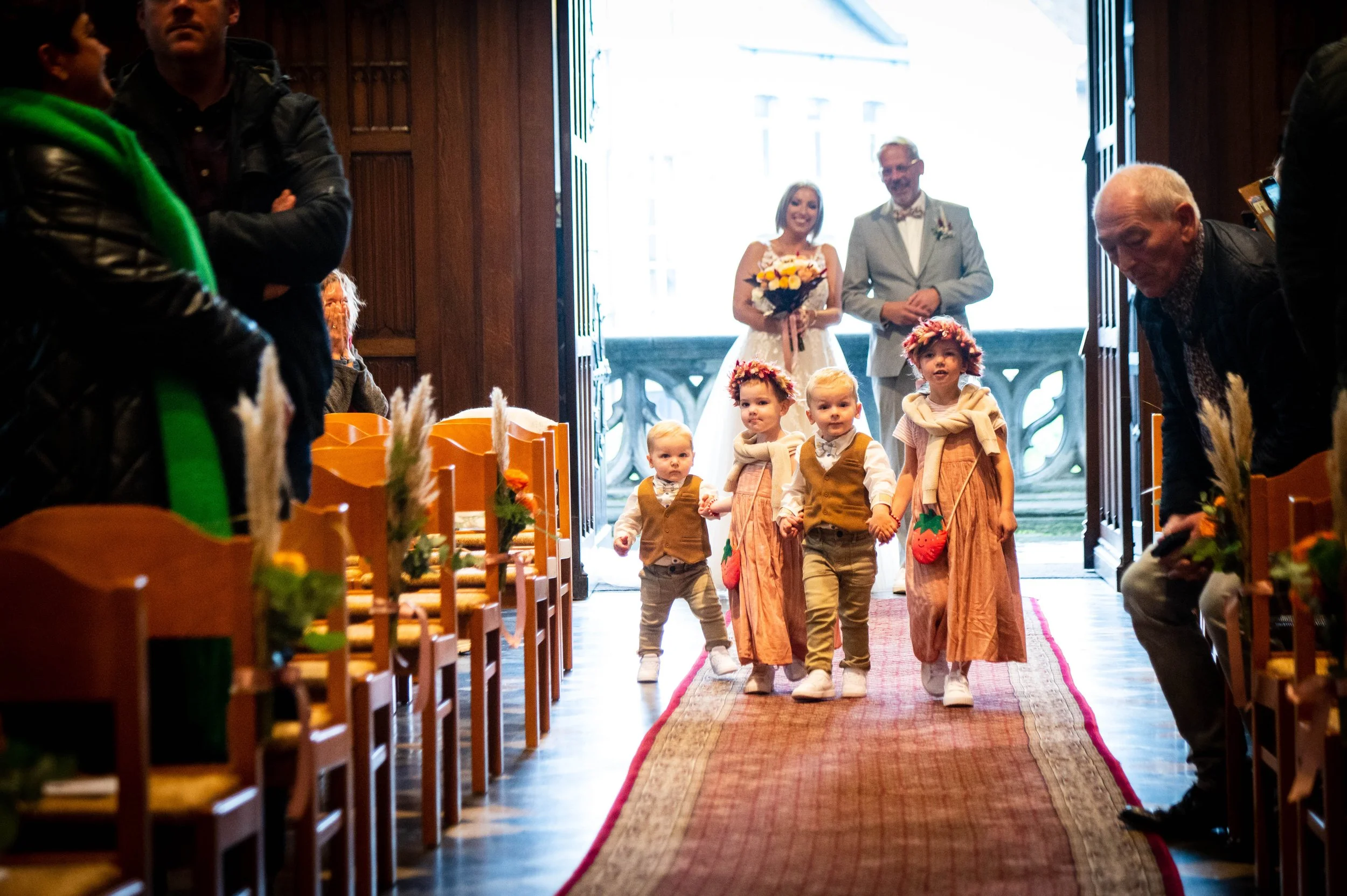 Een groep jonge kinderen loopt door de gang van een kerk of zaal, met een vrouw in een witte jurk en een man in een grijze pak die in de deuropening wachten. De kinderen dragen thee-omanen en bloemenkransen op hun hoofd.
