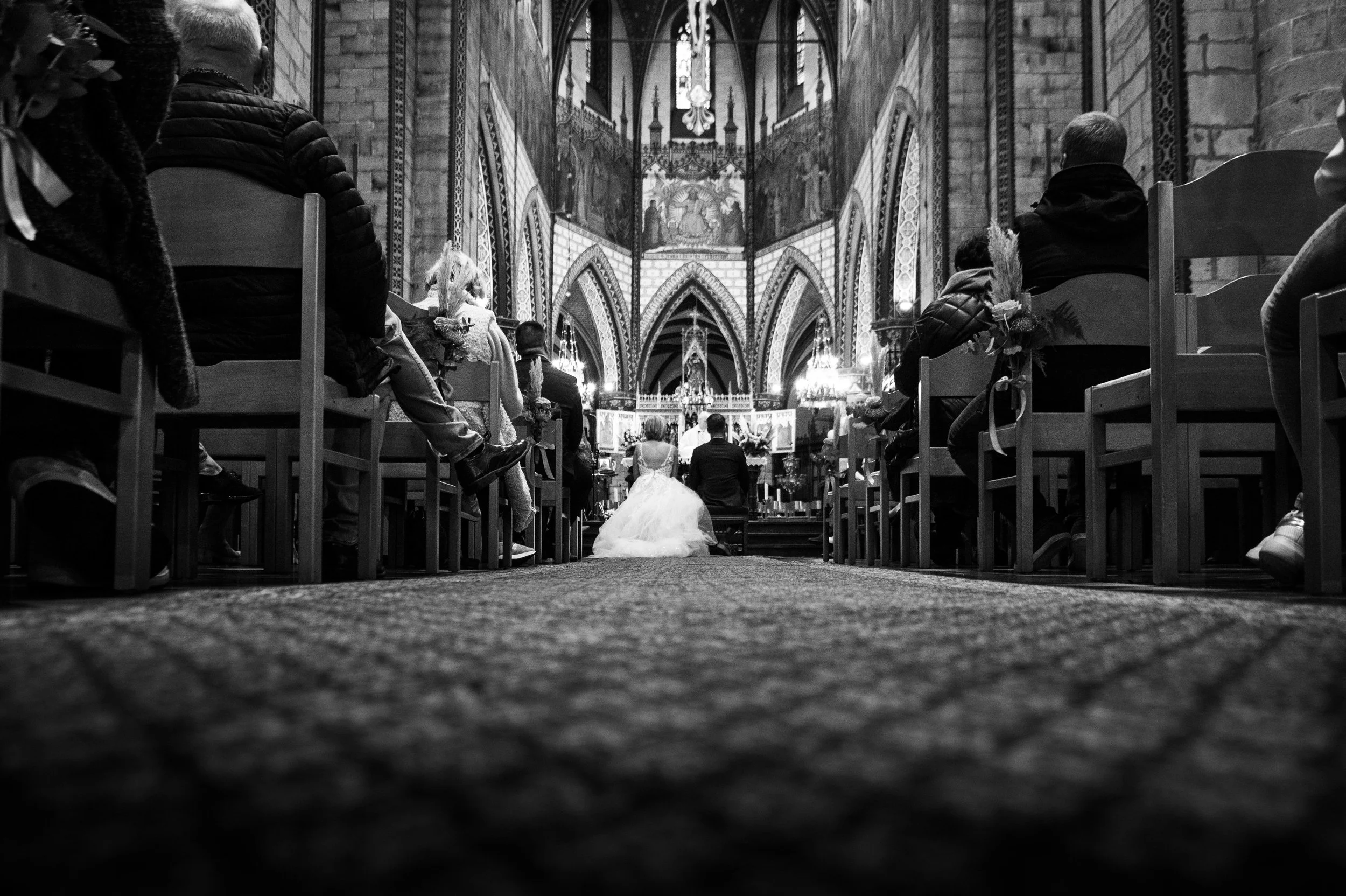 Bruidsparen zittend vooraan in een kerk tijdens een bruiloft. Fotografisch, zwart-wit, binnen, met publiek aan weerszijden van het gangpad, in detail en van een laag standpunt.