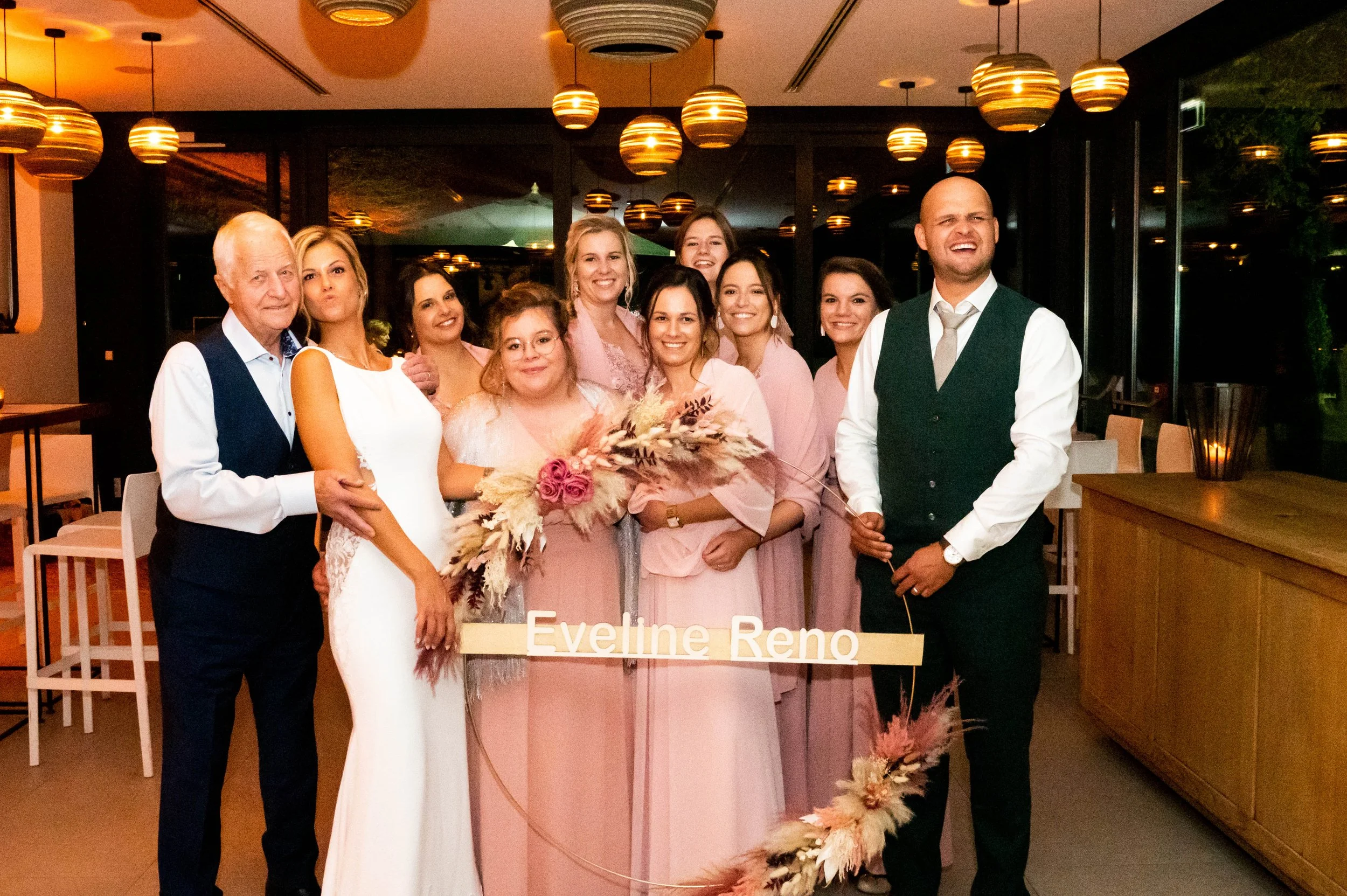 Een groep mensen op een bruiloft, met vrouwen in roze jurken en mannen in wit overhemd en zwart pak, inclusief het bruidspaar, binnen in een feestzaal met gedimde verlichting en ronde lampen aan het plafond.