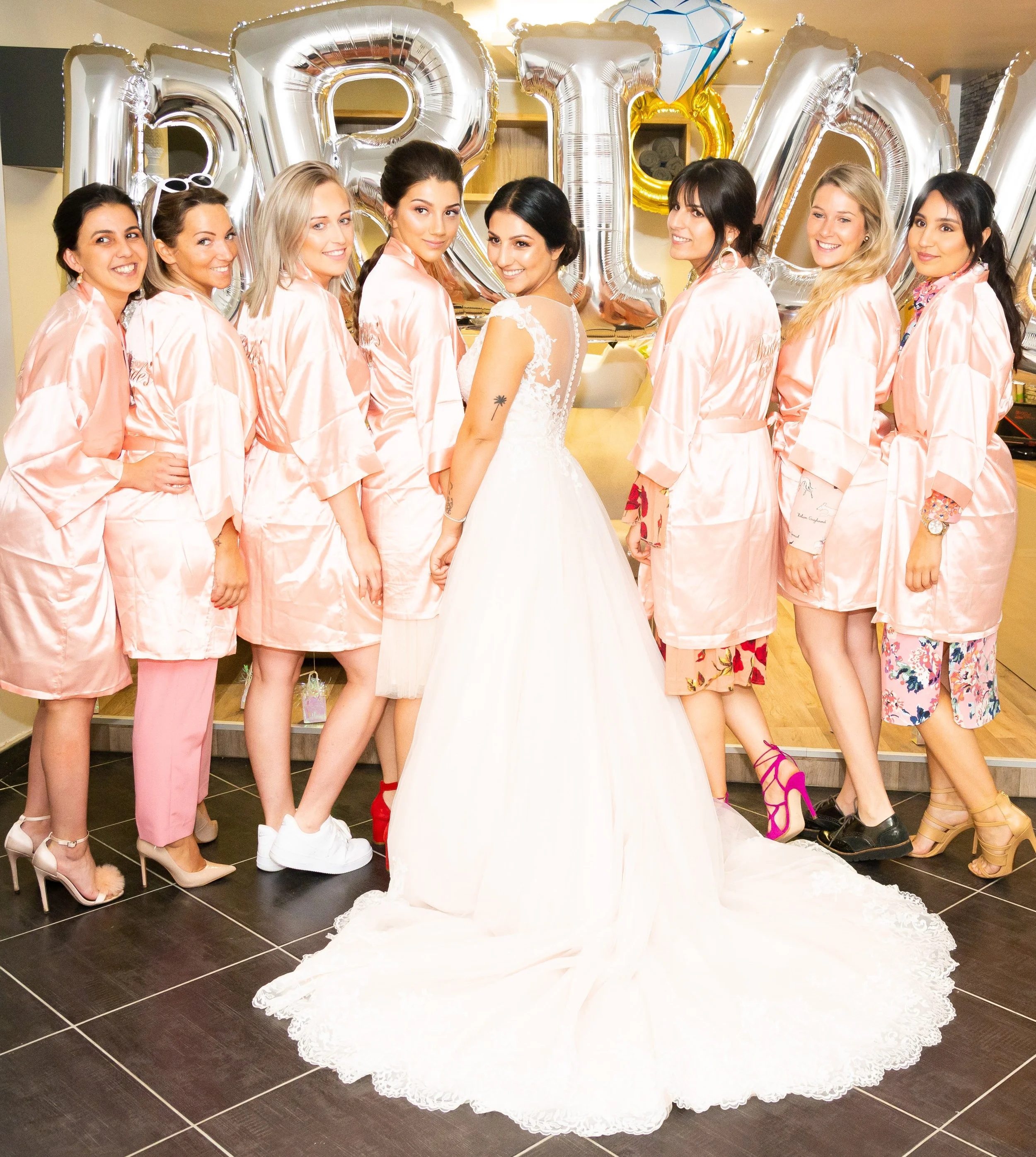 Een bruid in witte trouwjurk met haar vrienden of familie die allemaal in roze satijnen nachthemden poseren voor een foto, terwijl ze plezier maken en lachen tijdens een feest met grote zilveren en gouden ballonnen op de achtergrond.