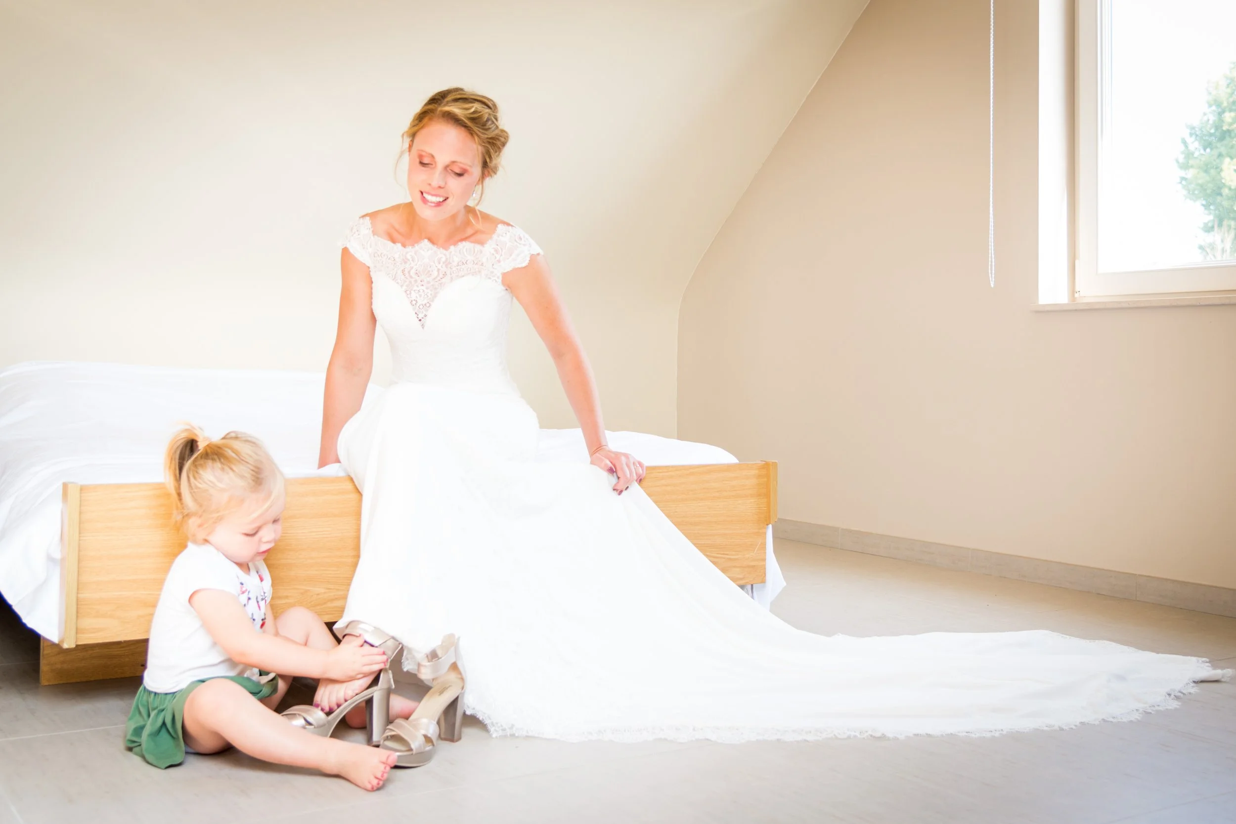 Een vrouw in een witte bruidsjurk helpt een jonge meisje met het aandoen van schoenen, in een lichte kamer met een bed en een raam.