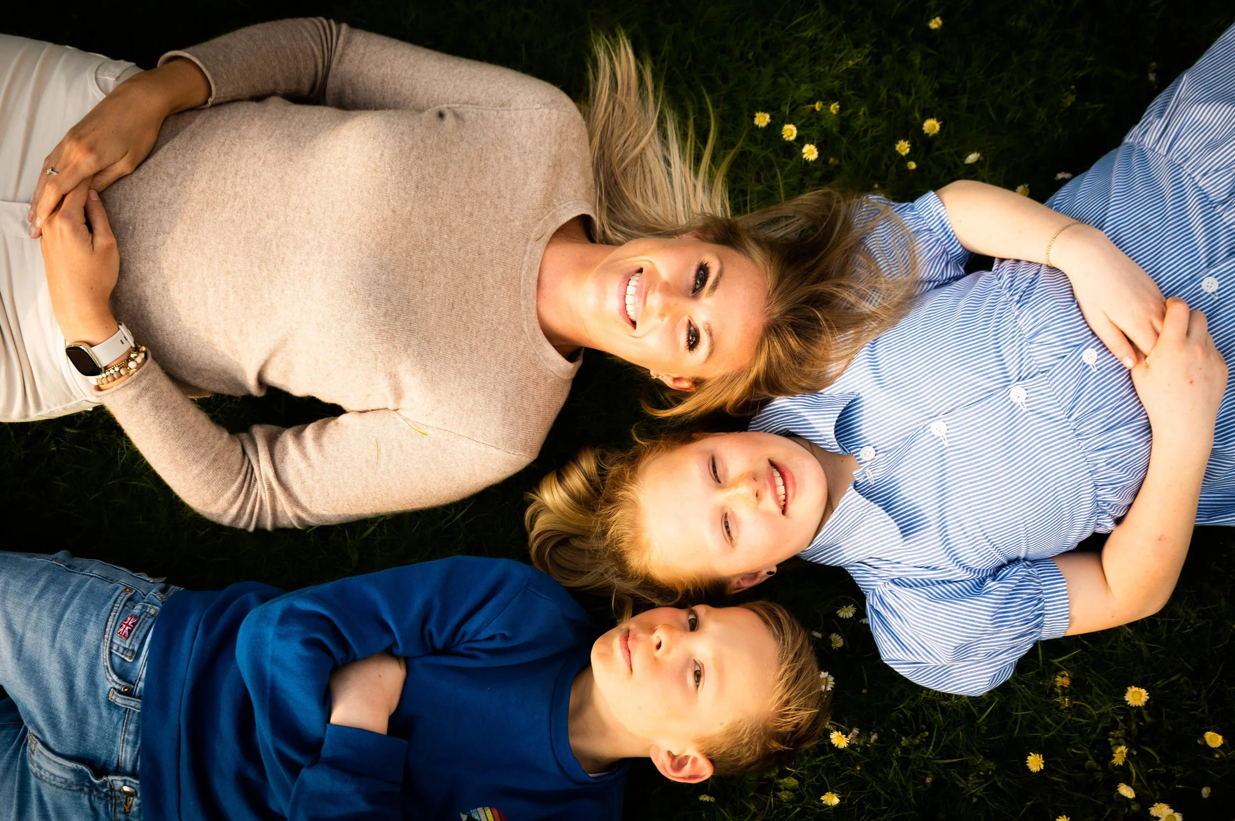 Vrouw met blonde haar ligt op gras met twee kinderen, een jongen en een meisje, alle drie glimlachend en ontspannen. Ze liggen op hun rug tussen kleine gele bloemen.