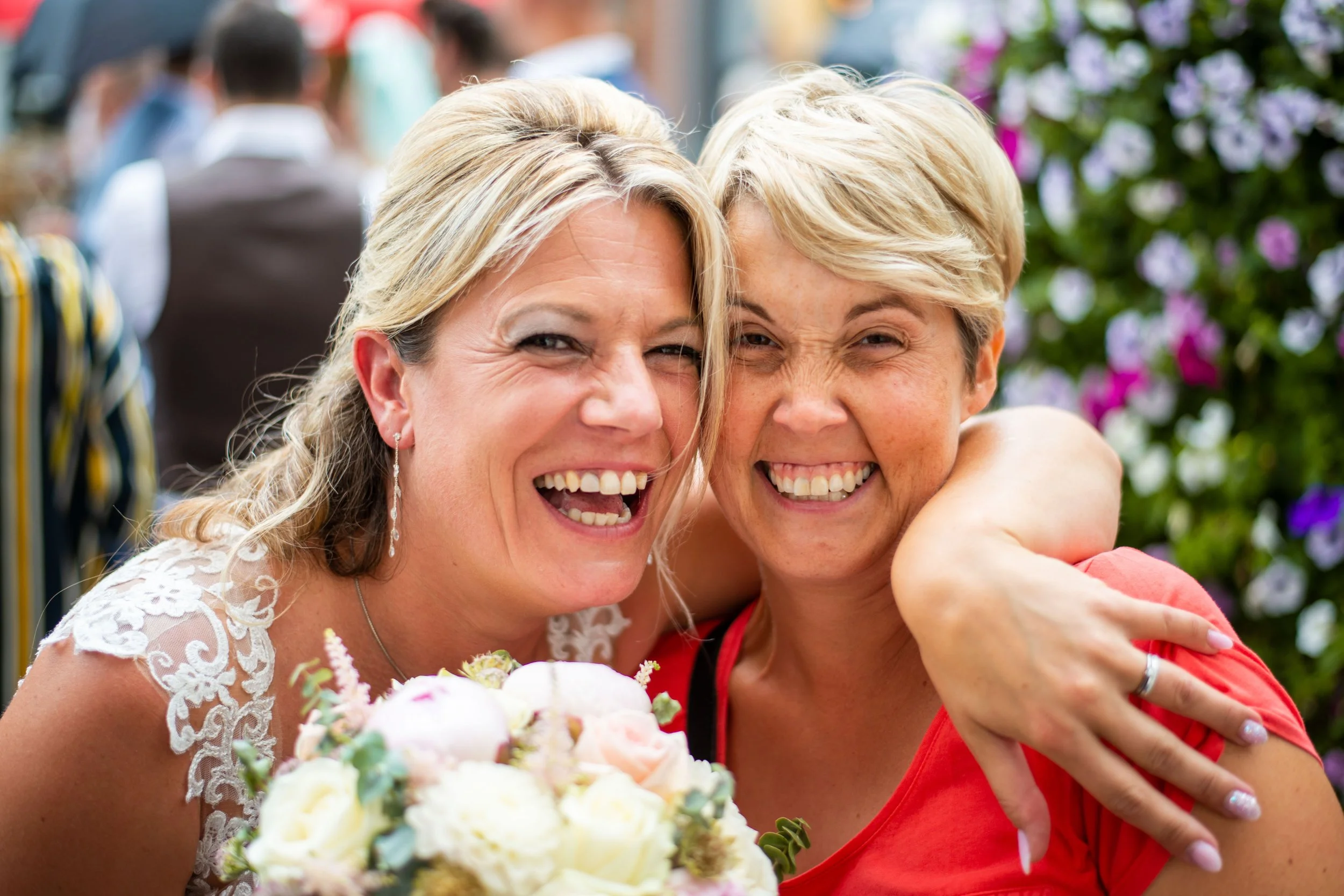 Twee vrouwen op een feest, lachen en omhelzen elkaar, met bloemen en mensen op de achtergrond.