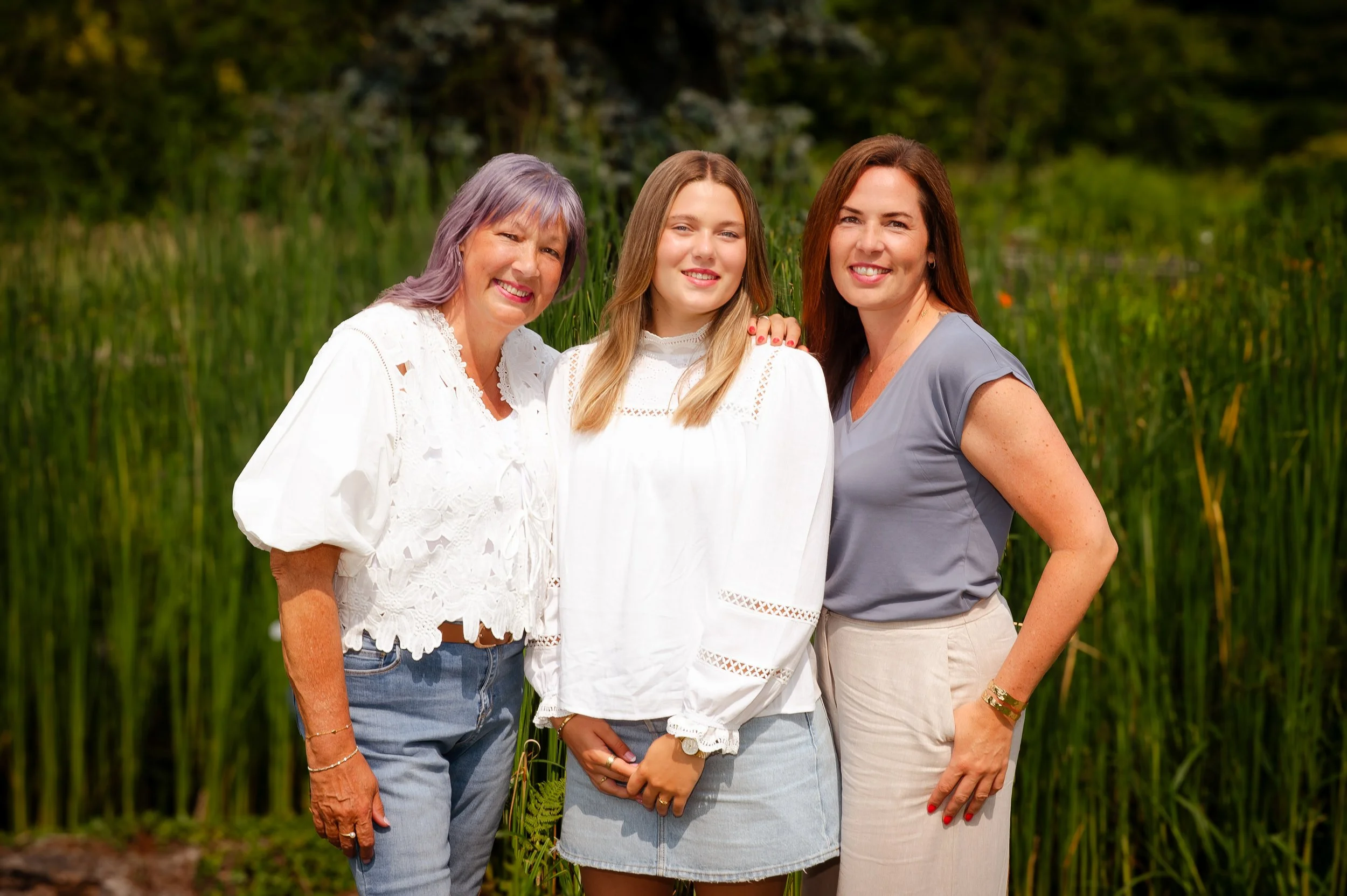 Drie vrouwen staan samen buiten in een groen landschap, lachen en kijken naar de camera, in zomerkleding.