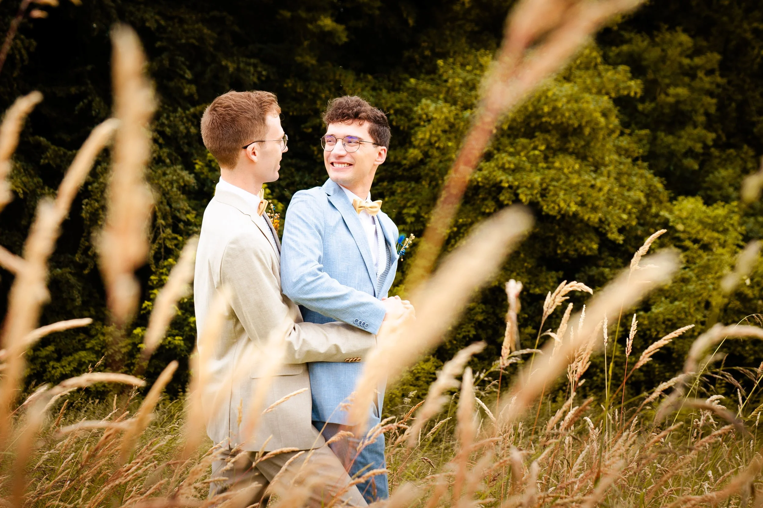 Twee mannen in pakken met strooien hoedjes die elkaars handen vasthouden en lachen in een veld met gras en bomen op de achtergrond.
