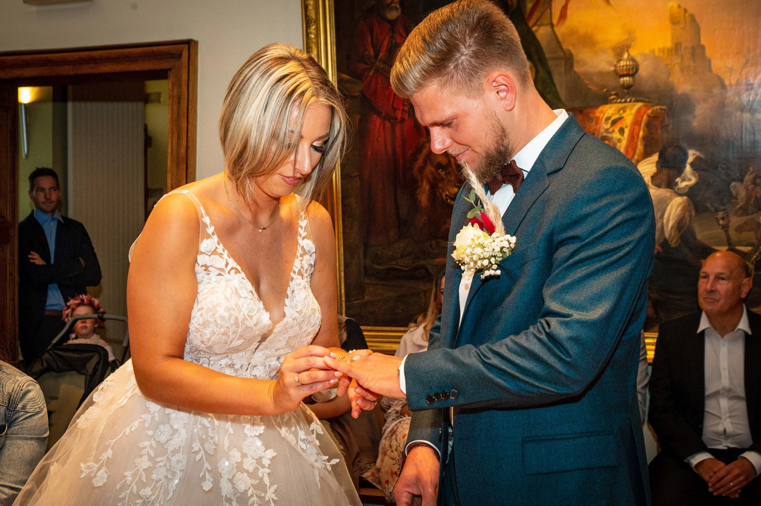 Een bruiloft waarbij een vrouw haar huwelijkssluiting in de ringvinger plaatst bij haar partner tijdens de ceremonie, met andere gasten op de achtergrond.