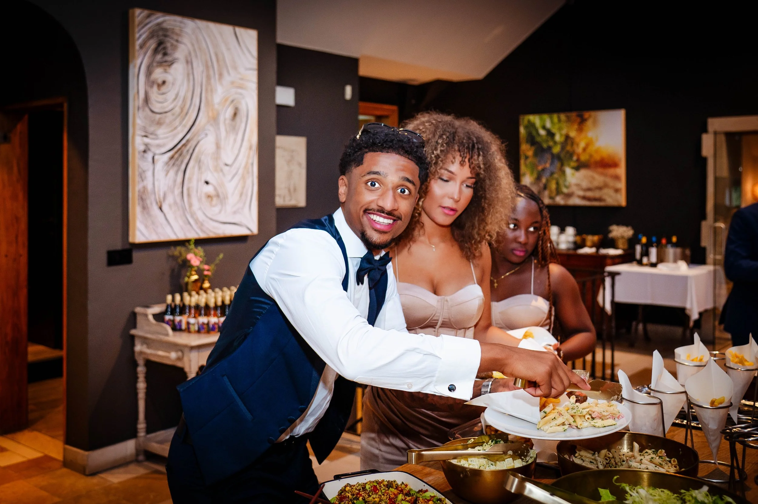 Man serveert salade en pasta tijdens een buffetfeest, terwijl twee vrouwen achter hem kijken.