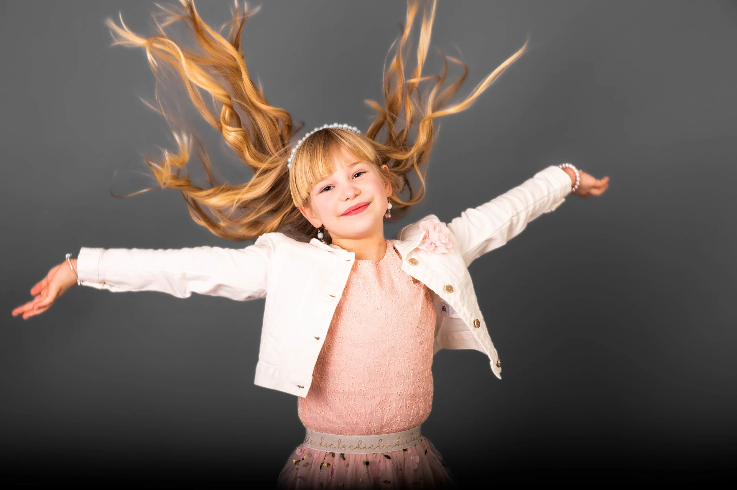 Meisje met lange, golvende blond haar die in de lucht hangt, lachend met uitgestrekte armen, tegen een grijze achtergrond.