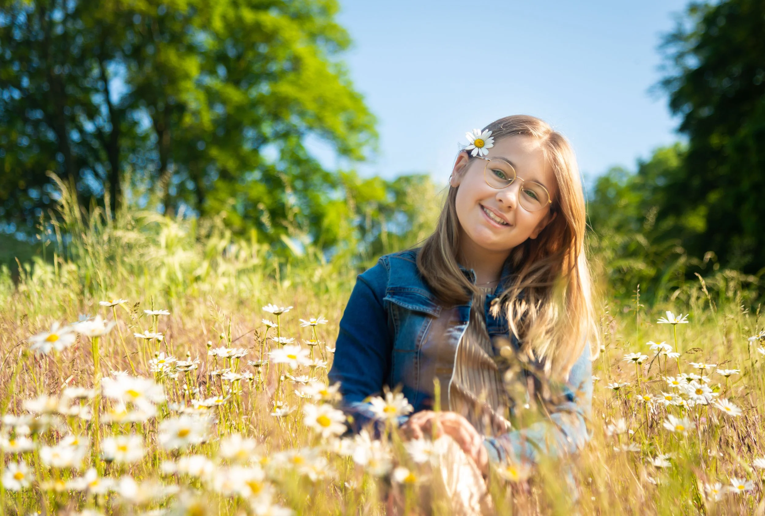 Vrouw met lang haar en een witte bloem in haar haar, glimlachend in een veld met madeliefjes onder een blauwe lucht, omgeven door groene bomen.