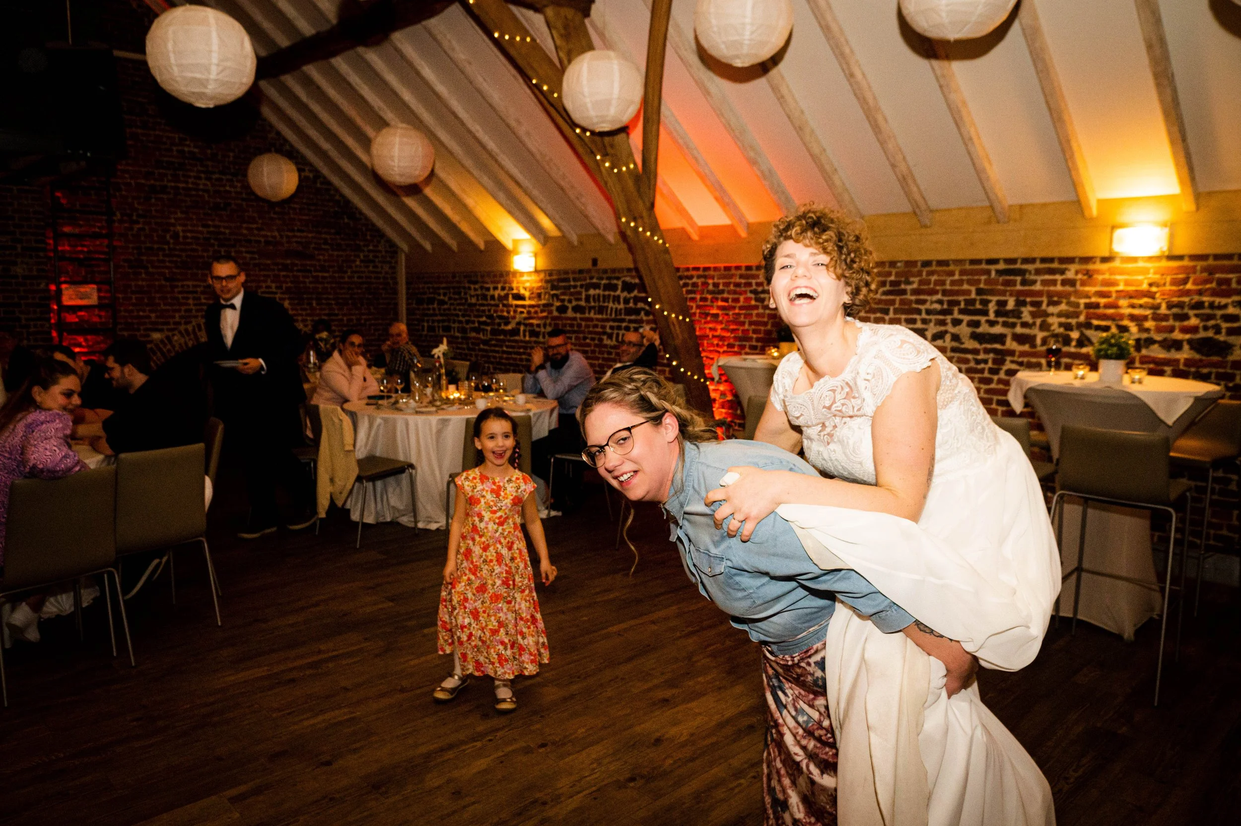 Vrouw in witte jurk die op de rug van een andere vrouw met bril staat en lacht, twee kinderen staan erbij, en mensen zitten aan tafels in een feestzaal met houten plafond en papieren lantaarns.