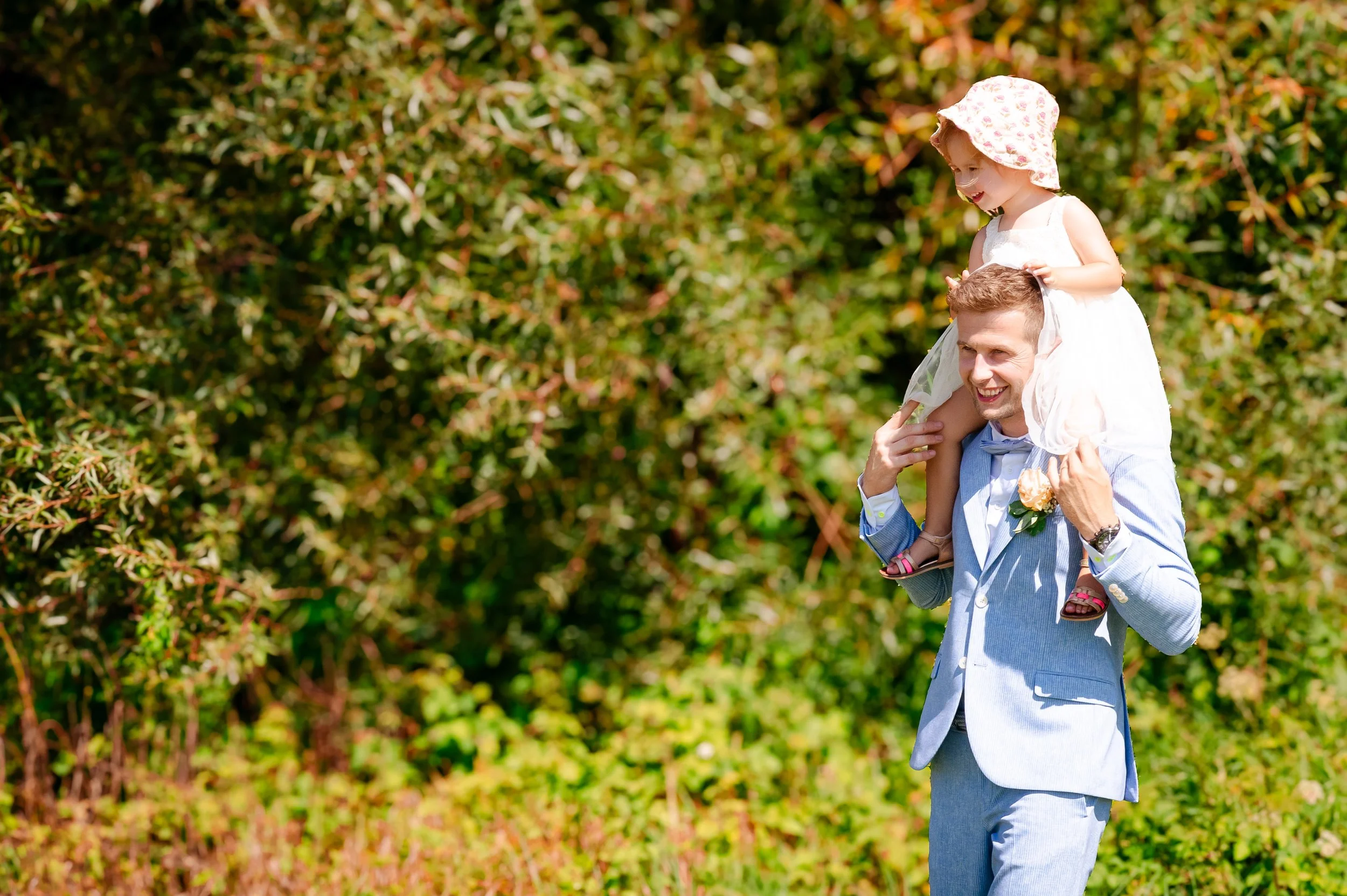 Man met een lichtblauw pak draagt een jong meisje in een witte jurk op zijn schouders, beide glimlachen en genieten van een zonnige dag in een groene buitenomgeving.