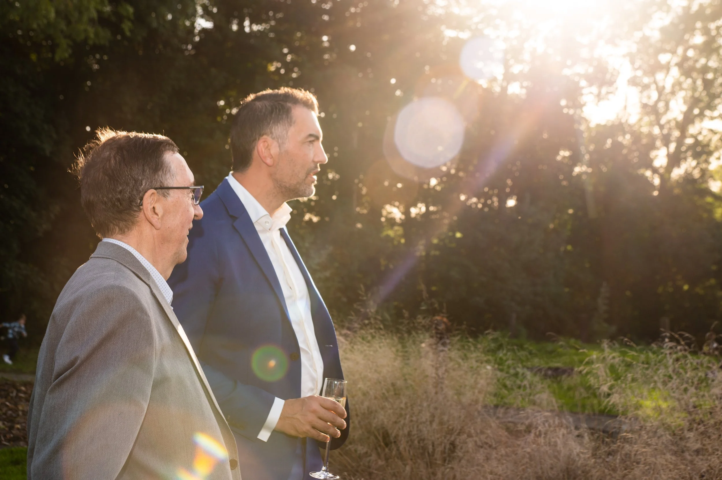Twee mannen in formele kleding staan in de buitenlucht tijdens zonsondergang, één houdt een glas champagne vast.