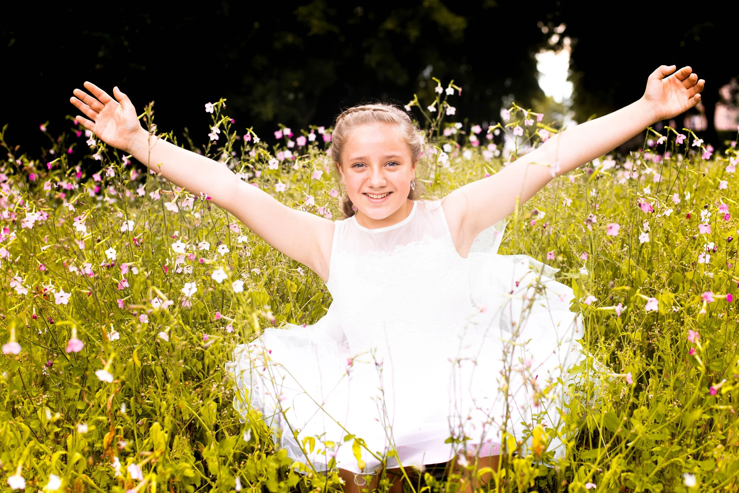 Vrouw in een witte jurk die in een veld met bloemen staat met uitgestrekte armen en glimlacht.