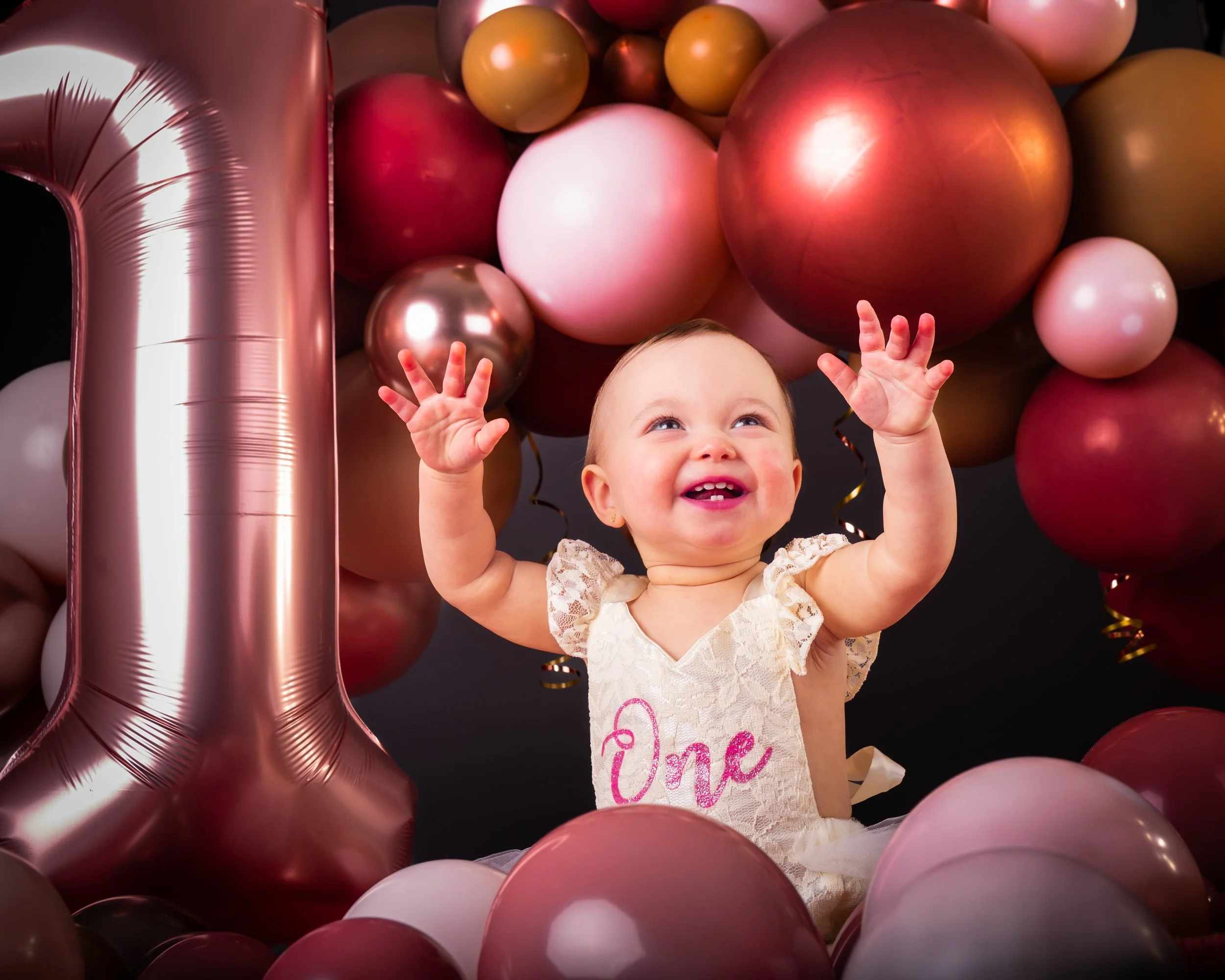 Gelukkige baby met jurk met 'One' tekst, omringd door roze, goud en witte ballonnen, met grote roze '1' ballon, vierkante zwarte achtergrond.