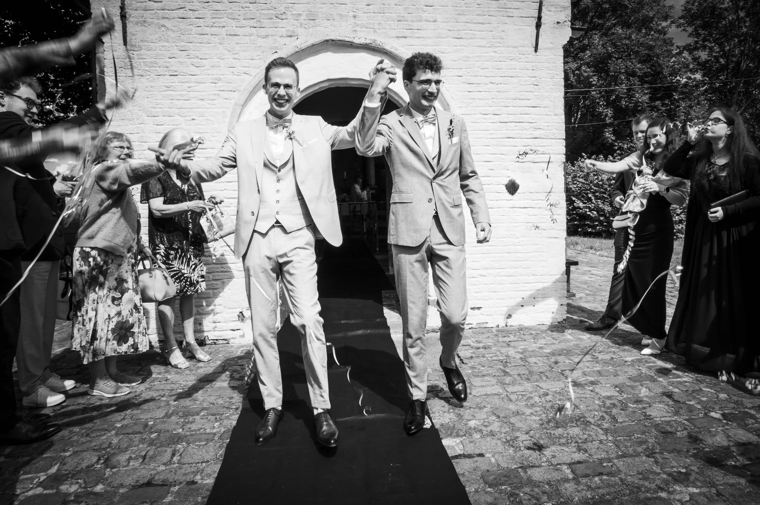Twee mannen in pakken vieren hun huwelijk buiten, omringd door gasten die applaudisseren en vieren.