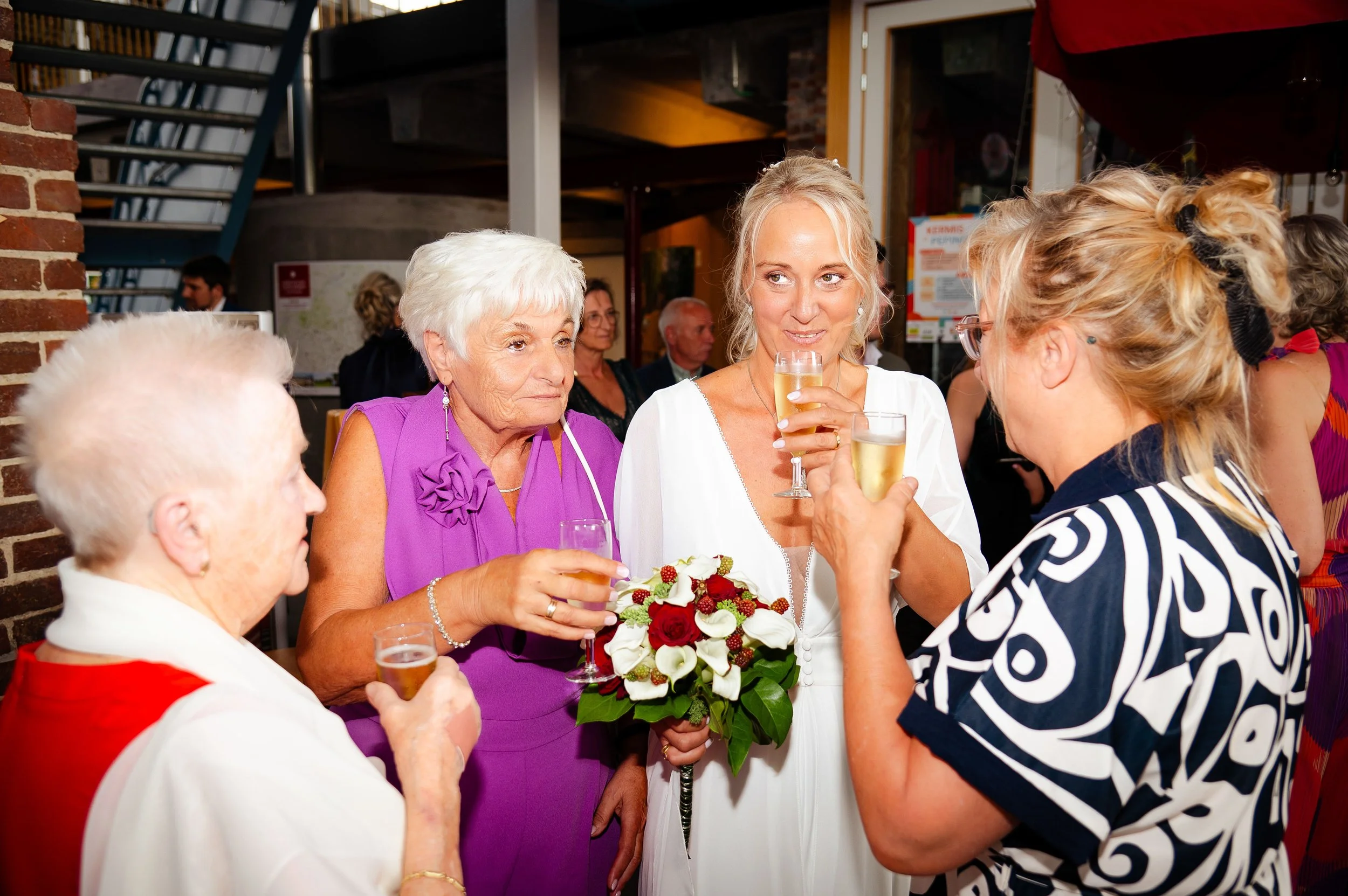 Vrouw met witte jurk en een boeket bloemen spreekt met drie oudere vrouwen tijdens een feest.