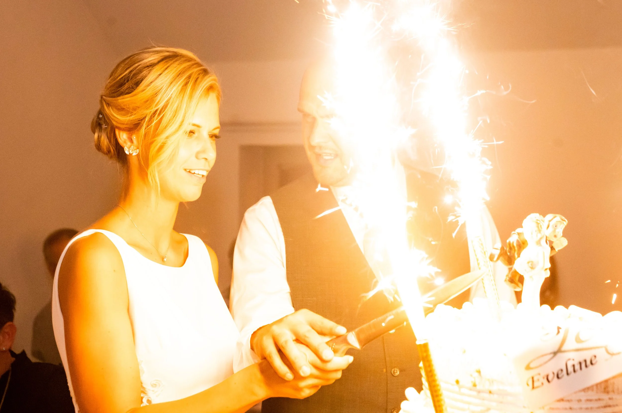 Een vrouw en een man vieren een verjaardag met een taart en een vuurwerkbrander, terwijl ze glimlachen en kijken naar het vuurwerk.