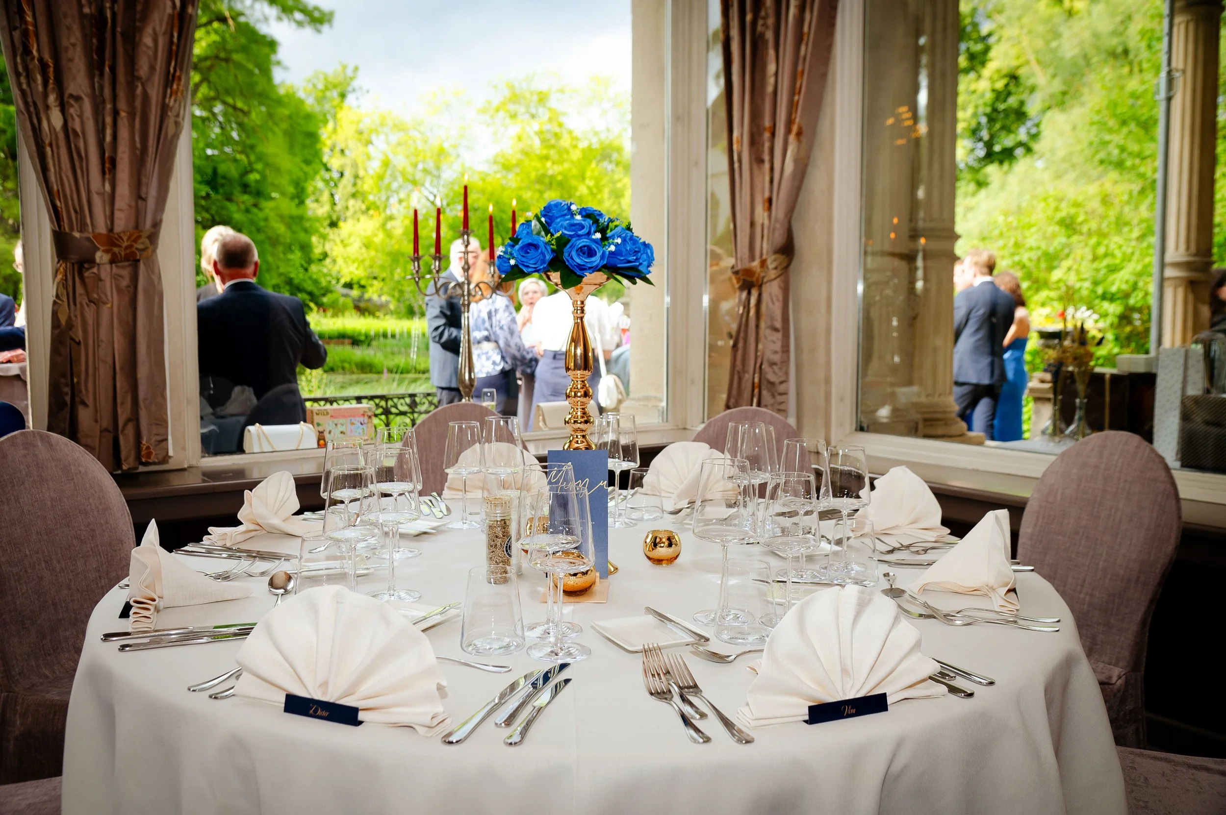 Elegante gedekte tafel in een feestzaal met uitzicht op een tuin. Centrale bloemstuk met blauwe rozen en gouden kandelaars, omringd door glazen en bestek.