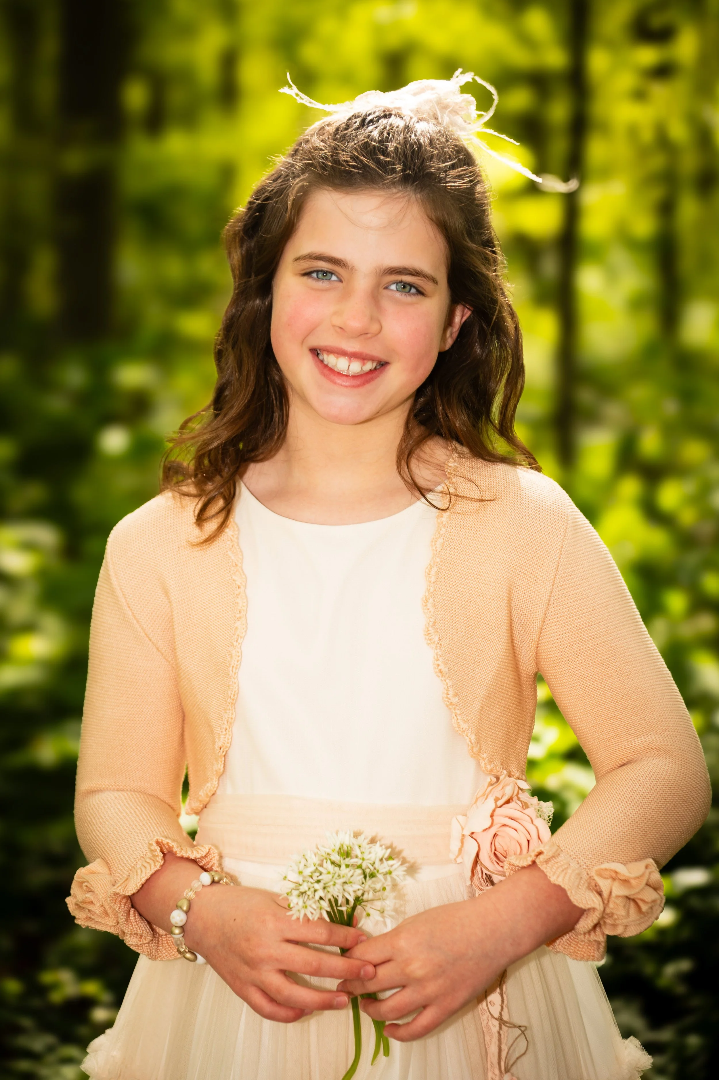 Een jonge meid in een lichte jurk en een roze jas met franje, die in een bos staat en een boeket kleine witte bloemen vasthoudt. Ze glimlacht naar de camera.