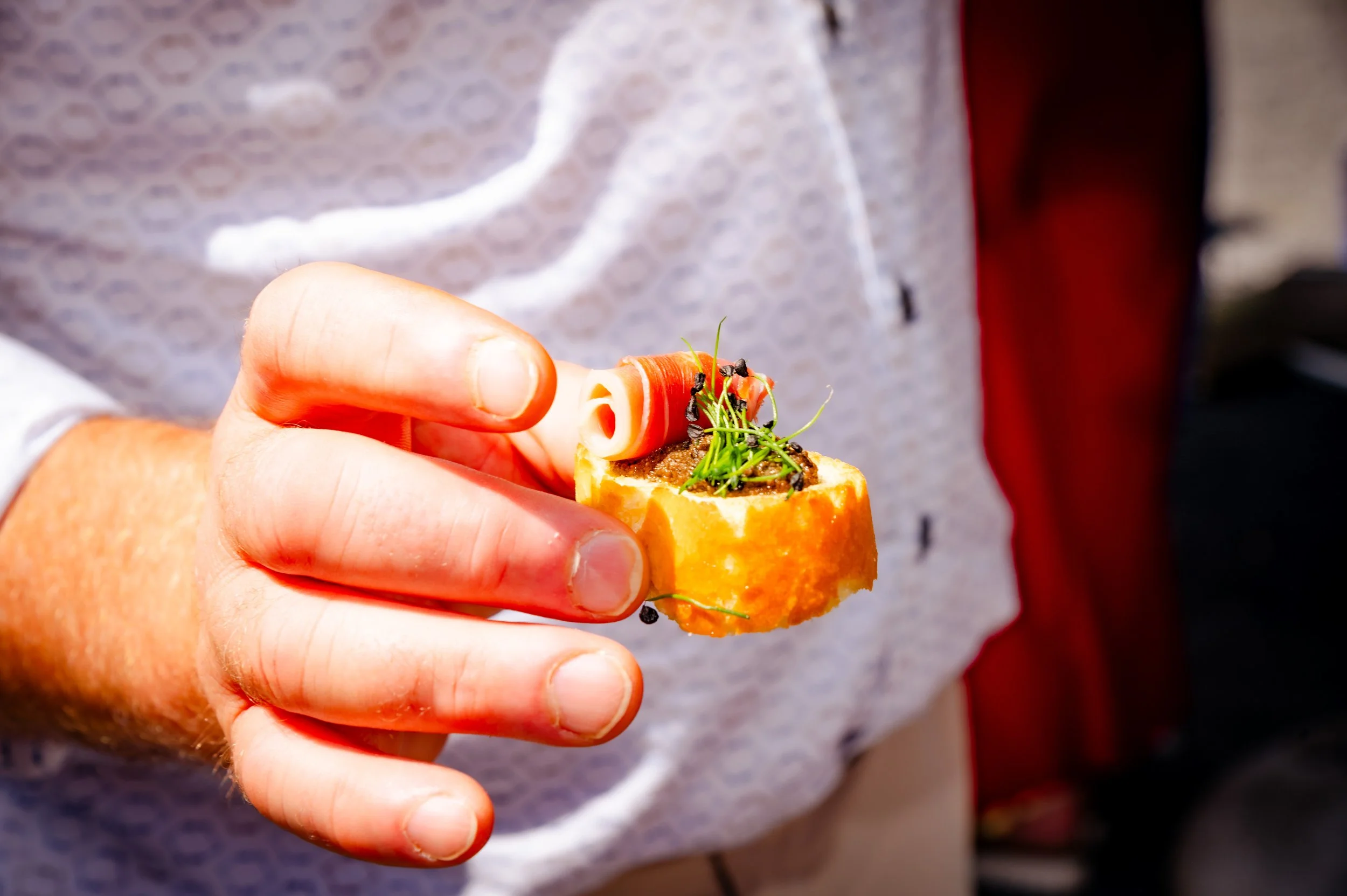 Close-up van een hand die een kleine canapé met ham, imitatiekaviaar en microgroenten vasthoudt.