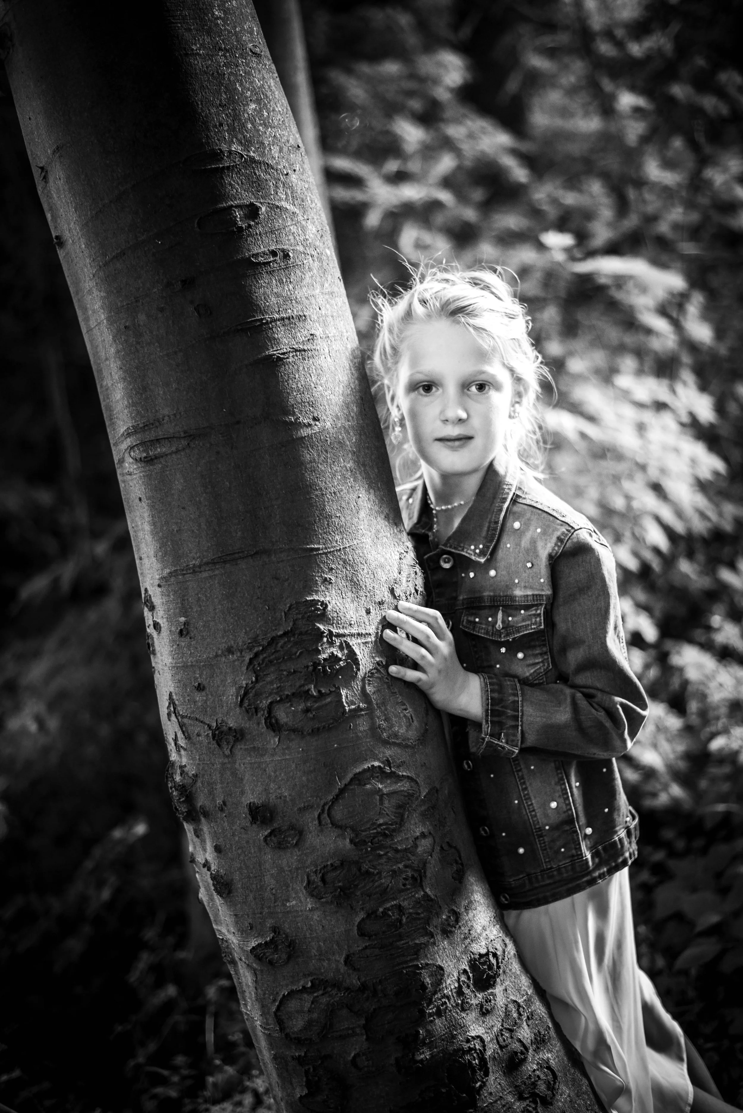 Jonge meisje met blond haar die zich achter een boom verschuilt in een bos, zwart-wit foto.