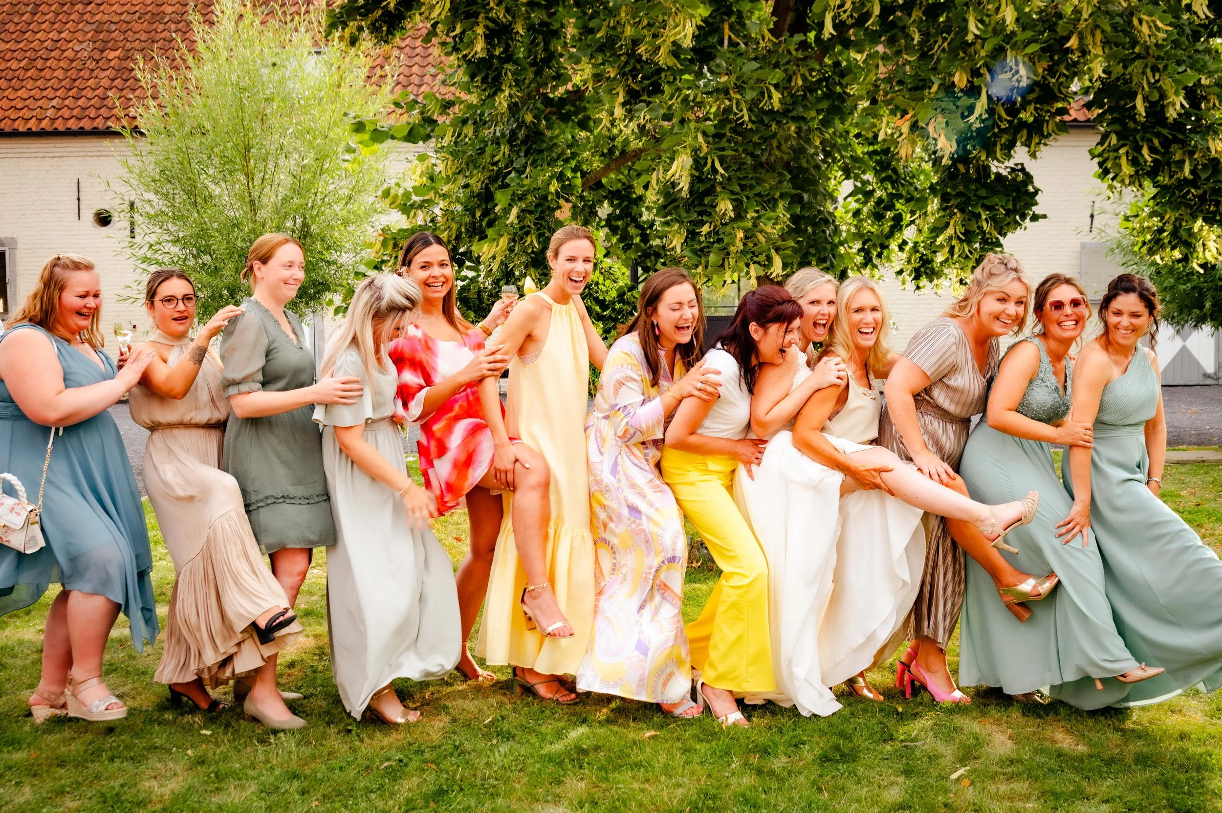 Glimlachende vrouwen in kleurrijke jurkjes die samen optrekken, met een groene tuin en boerderijgebouwen op de achtergrond.