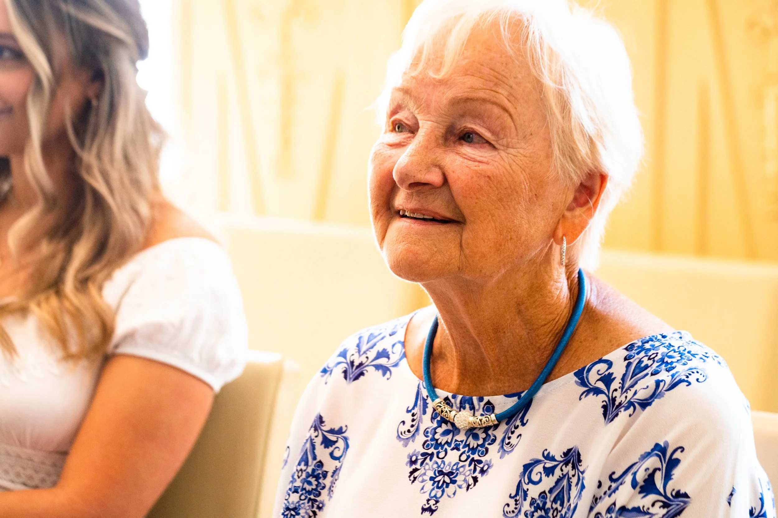 Een oudere vrouw met wit haar die lacht, zittend aan een tafel, draag een wit blouse met blauwe patronen en een blauwe ketting, op de achtergrond een lichte muur.