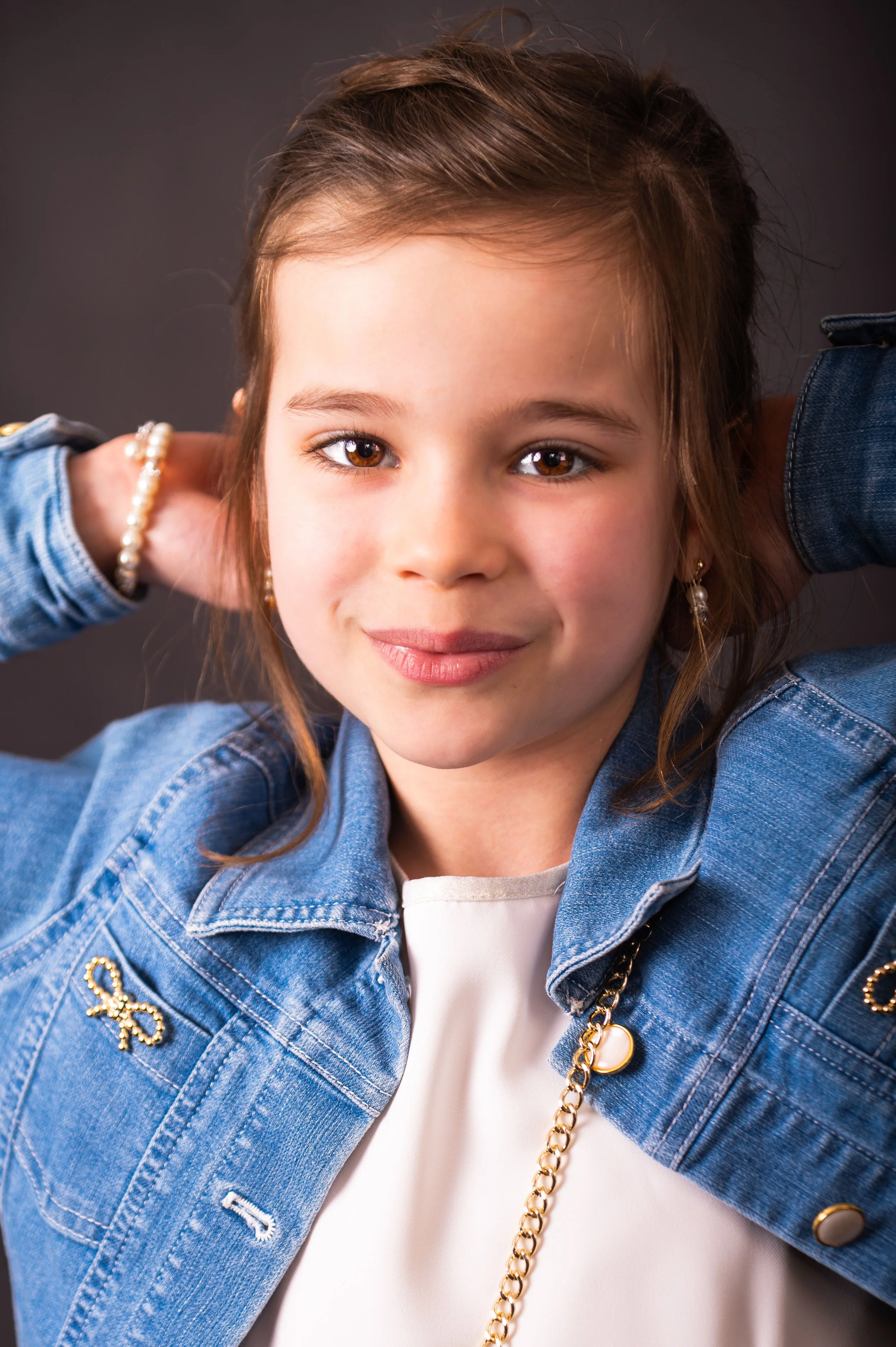 Portret van een jonge meisje met bruin haar, die glimlacht en haar handen achter haar hoofd houdt, gekleed in een denim jas en witte top.