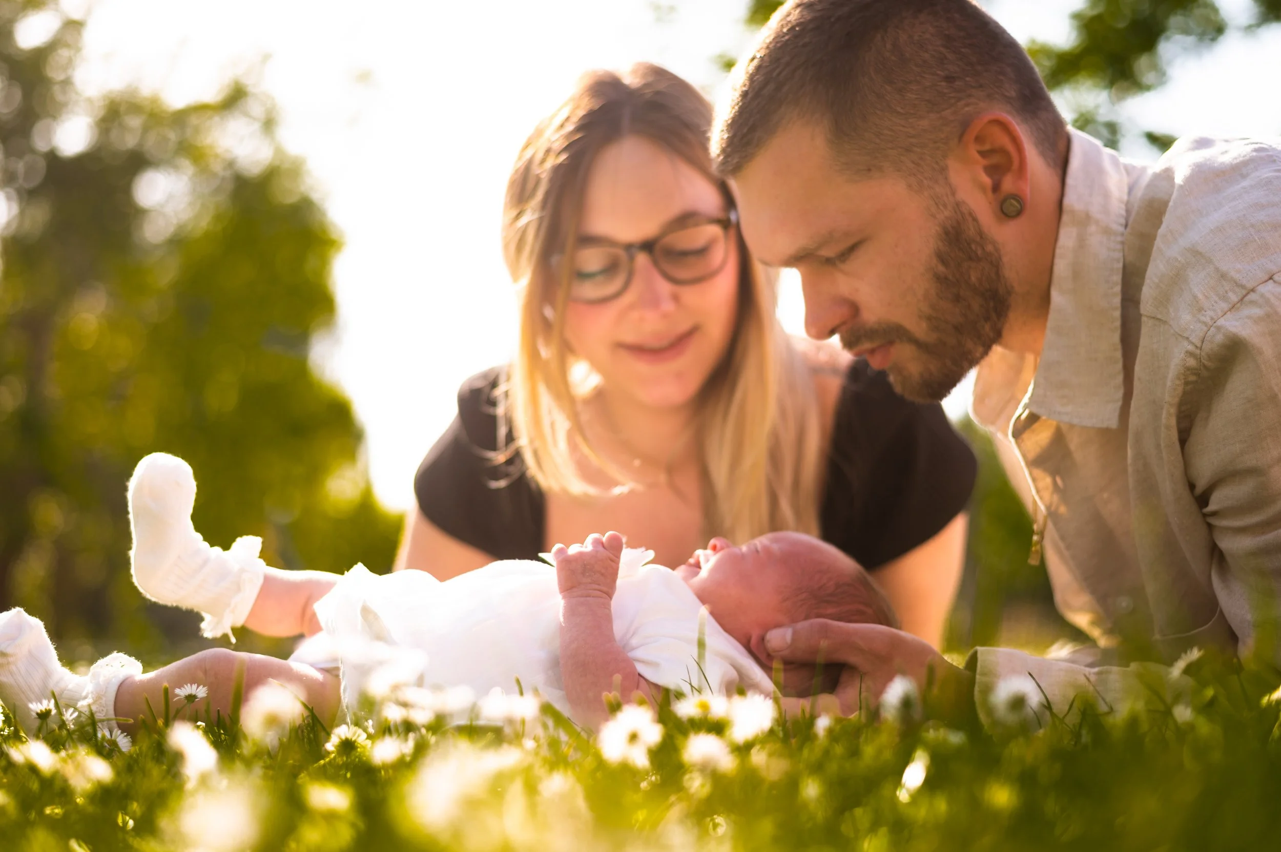 Een jonge familie ligt op een grasveld met kleine witte bloemen, nakend in de zon, met een moeder, vader en baby.