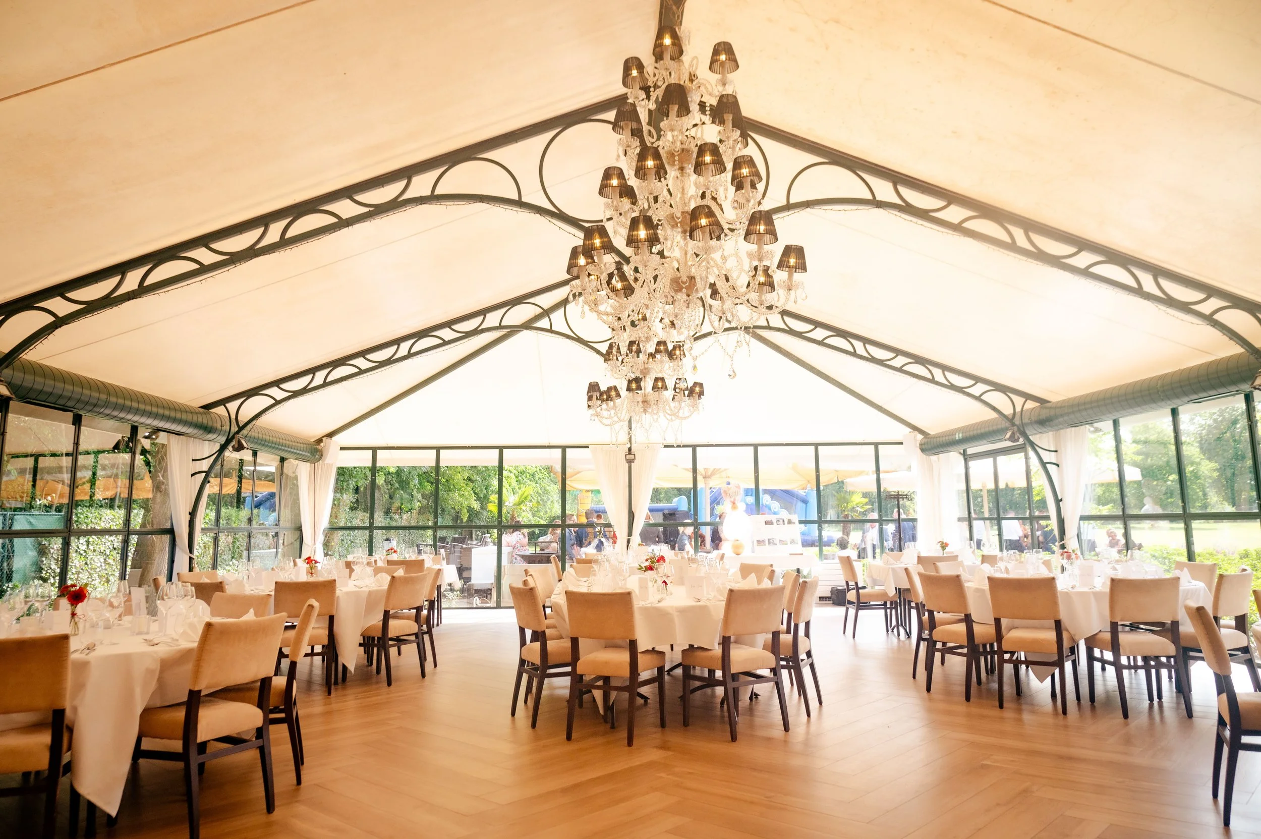 Elegante feestzaal met ronde tafels bedekt met witte tafelkleedjes, versierd met bloemen, en beige stoelen, grote glazen muur met uitzicht op tuin, zware chandelier aan plafond.